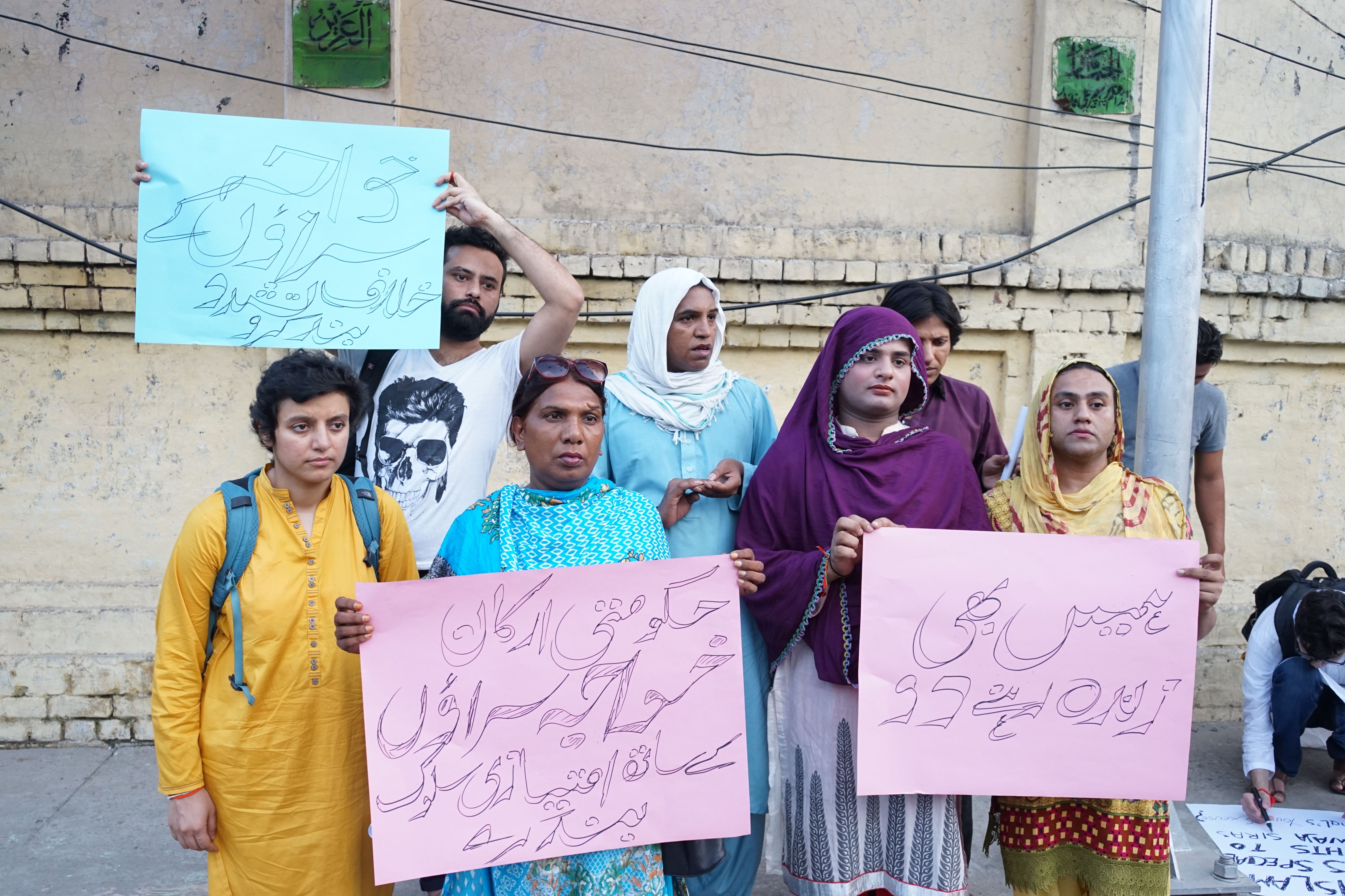 A group of protesters stand with signs on the sidewalk of a busy street. It is near evening in early August. The blue sign reads, "Stop the violence against khawaja siras." (The other two signs are the same from the previous picture). Image by Ikra Javed. Pakistan, 2016.