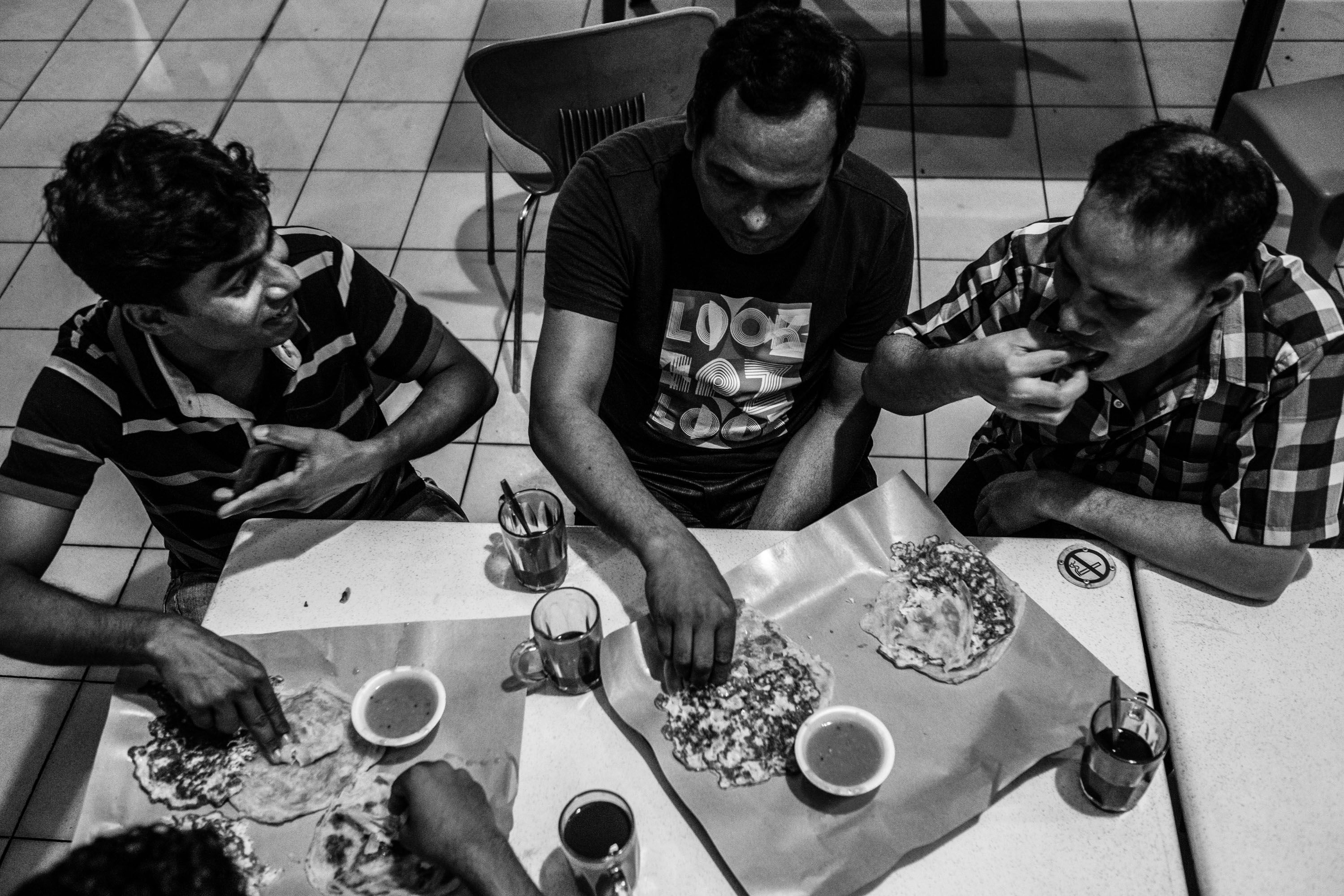 Mr. Haque and his friends enjoy a simple meal in Singapore. Roti prata, a fried flat bread, is their meal of choice because it is cheap. Image by Xyza Bacani. Singapore, 2016.