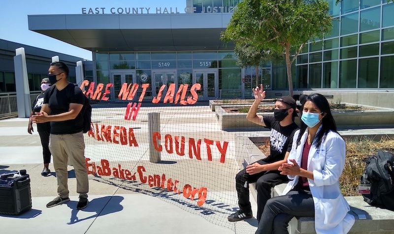Three protestors stand outside Santa Rita Jail in front of a sign that says 'Care not jails in Alameda County - ellabakercenter.org.' Image by Andrew Stelzer / KALW. United States, undated.