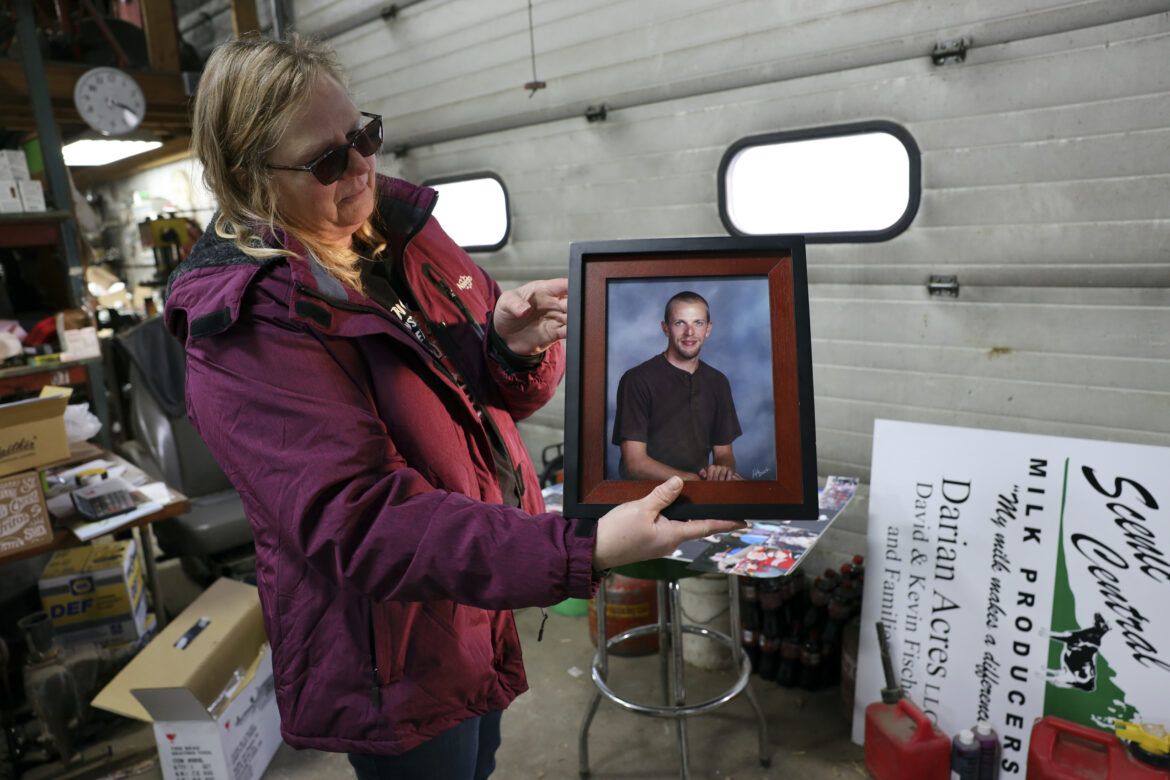 Amy Fischer is seen with a photo of her son, Brian, on her family’s 350-cow dairy farm, Darian Acres, in Rio, Wis., on Dec. 18, 2020. Brian died by suicide at the age of 33, on Dec. 21, 2016. The Fischers attribute his death to a combination of stress from work, a drinking problem and depression from a recent break-up. Dairy farmers and their advocates say numerous stressors are leading to a mental health crisis in their industry, including financial pressures, long hours, labor shortages and harsh weather. Image by Coburn Dukehart / Wisconsin Watch. United States, 2020.