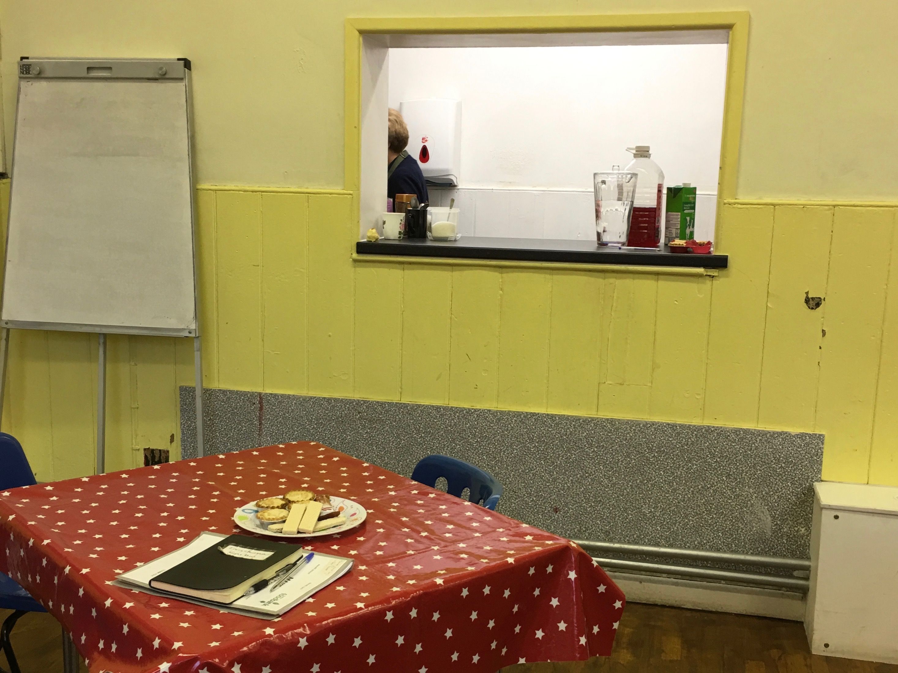 The peep hole to a small kitchen. Coffee and tea are always available for the clients, as well as biscuits and treats on the table. There is also a guest book which encourages clients to write down their stories. These are then used to end stigma around individuals using food banks. Image by Caitlin Bawn. England, 2016.