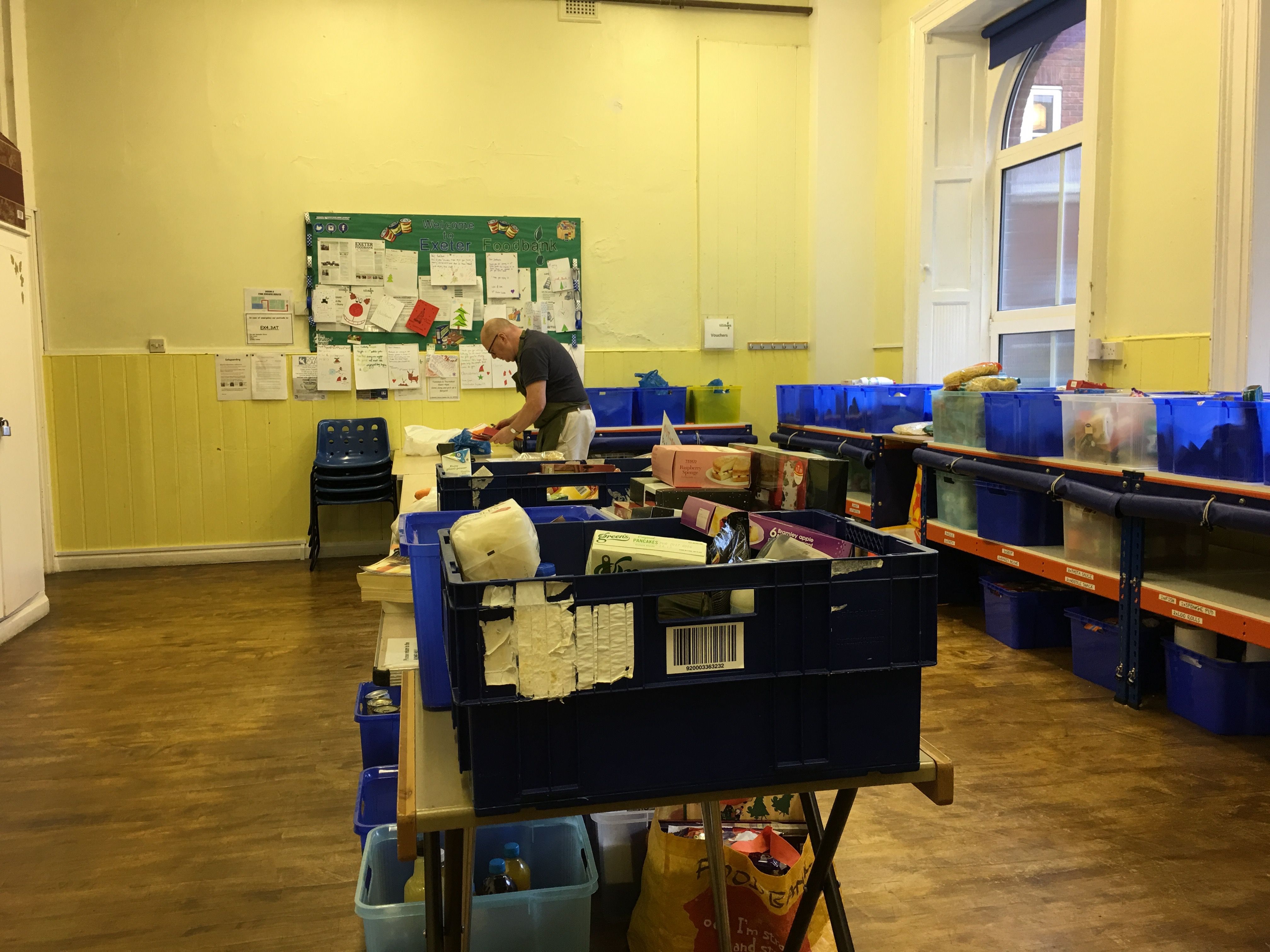 A volunteer clears up after the afternoon session, putting the unused food back into the cupboards lining the wall. The church hall offers highly versatile space - useful and necessary for this expanding organisation. Image by Caitlin Bawn. England, 2016.