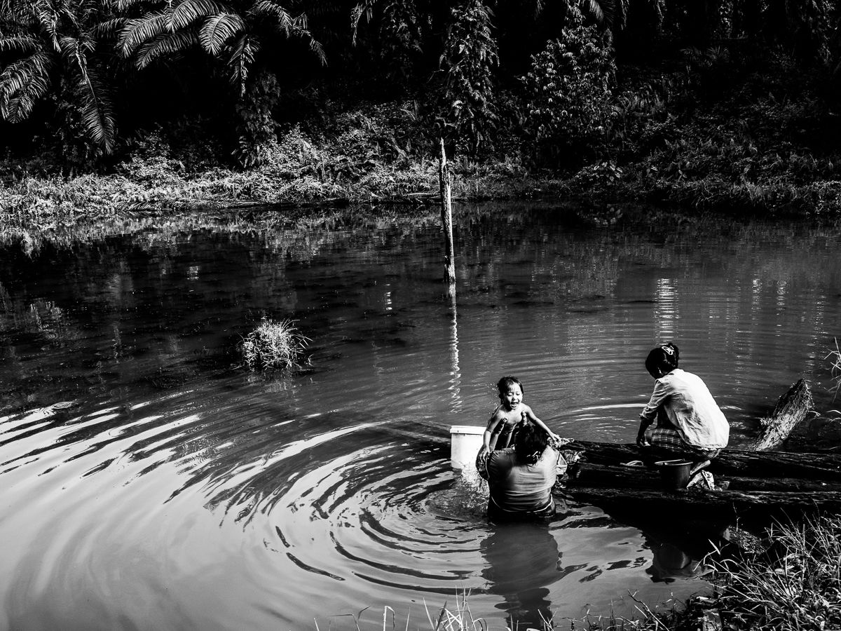 WATER SOURCE. Santi takes a bath in the murky water with a neighbor while her mother Ida washes their clothes. The reservoir is the community's only source of water. Image by Xyza Cruz Bacani. Indonesia, 2018. 