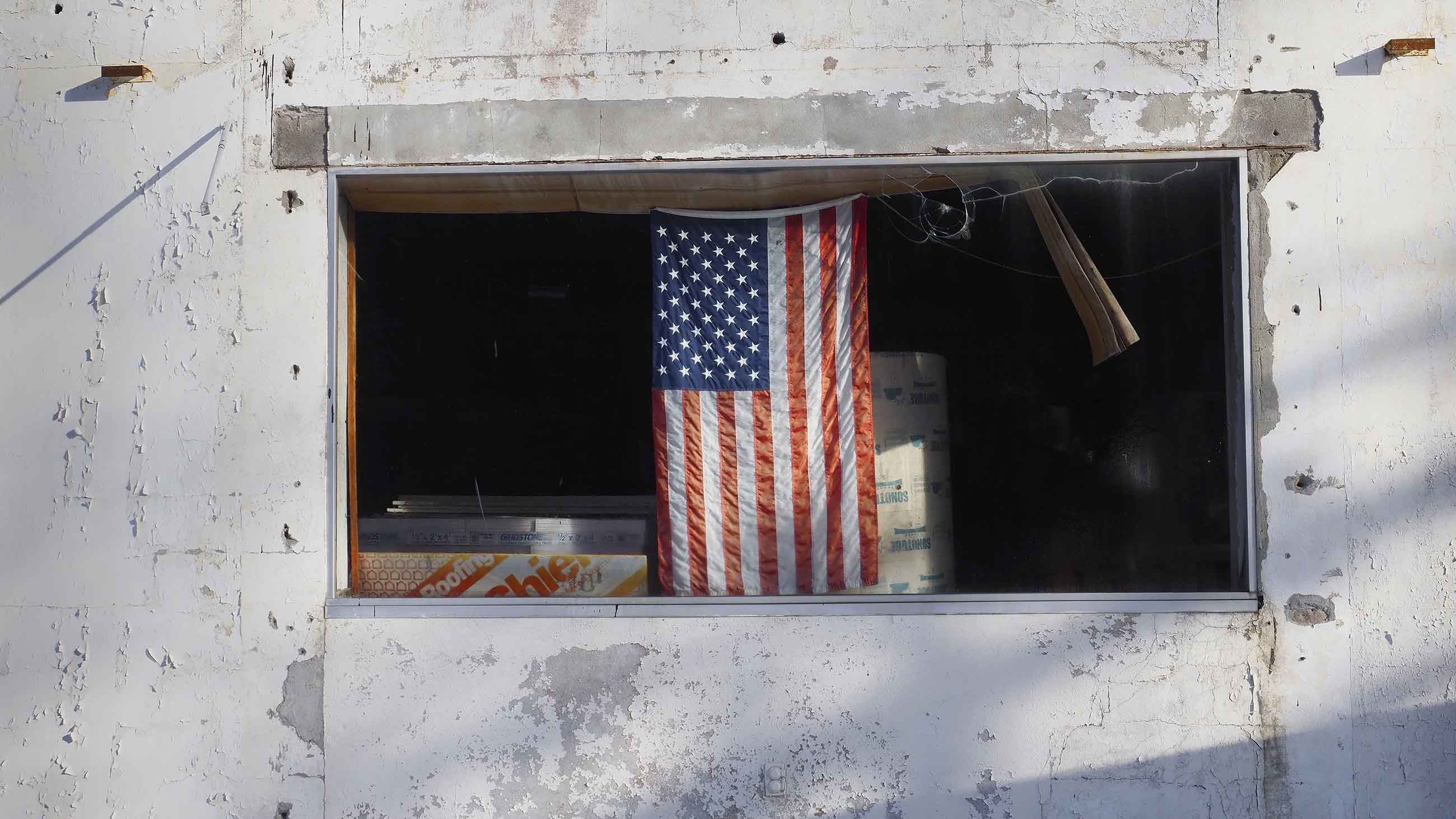 A flag hangs in the window of a small building on the grounds of the former Independent Leather Manufacturing Corporation in Gloversville, one of dozens of Superfund and brownfield sites in and around town. Image by Larry C. Price. United States, 2016.