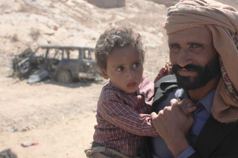 Mabkhout Ali al Ameri stands with his 18-month-old son, Mohammed, in the village of al Ghayil in Yemen’s al Bayda province. Mabkhout’s wife, Fatim Saleh Mohsen, was shot in the back of the head by helicopter gunship fire as she fled with Mohammed in her arms during a U.S. raid. Image courtesy of The Intercept. Yemen, 2017.