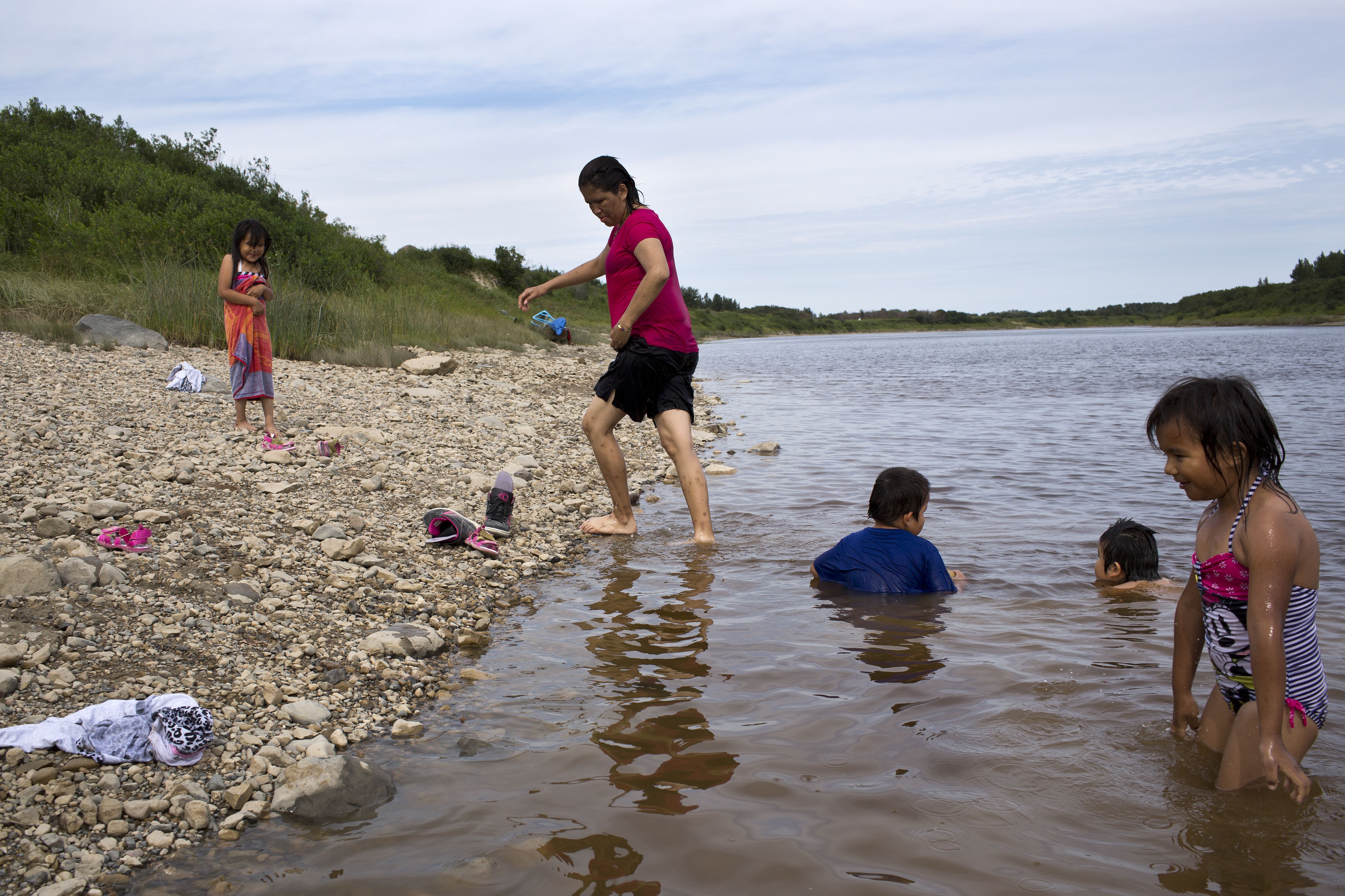 Twenty-seven-year-old Samantha Iahtail (centre, in red) swims with her nieces and nephews on the banks of the Attawpiskat river. Samantha tries to help her sister with the kids when she can and while her life has been filled with challenges of her own, she believes that "the creator doesn’t give you things you can’t handle”. In Attawapiskat many families face great difficulties yet respond with resilience, relying on each other to navigate the struggles associated with being First Nations people. Image by David Maurice Smith. Canada, 2016.