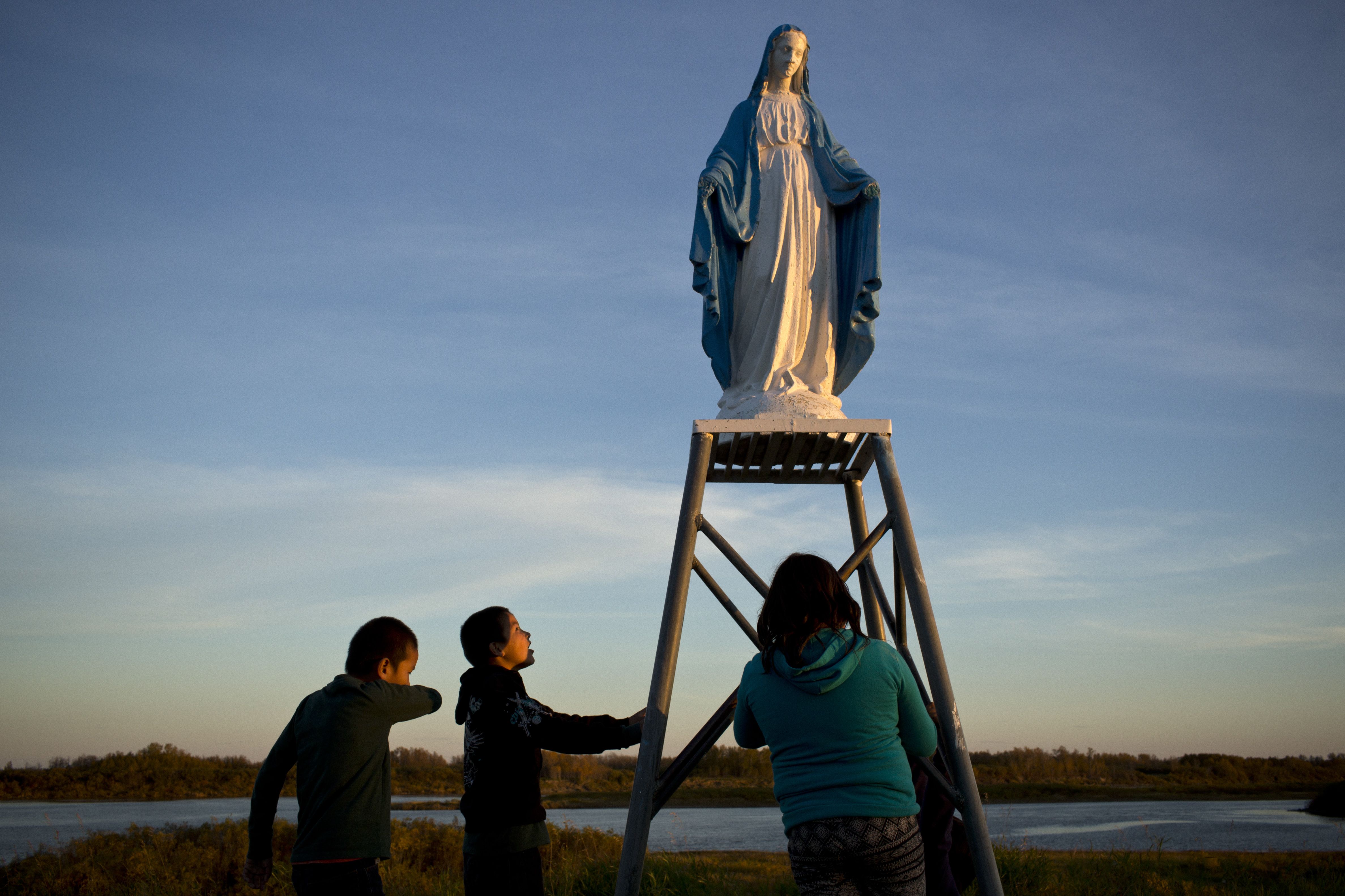 Young people play at a statue of the Virgin Mary near the St. Francois-Xavier Catholic Church on the shores of the Attawapiskat river. The community has a complicated history with the Catholic Church, since many of its members were subjected to horrible abuses while at the Catholic-run St. Anne’s residential school. A conversation with one 59-year-old man who had endured St Anne's for six years remained with me; he told me of repeated sexual and physical abuse at the hands of priests and nuns and, in one particular instance, a nun punched him and broke his nose when he was 11. It was the last time in his life that he was able to cry, he said. To this day he still carries deep shame and wishes he could express it through tears—however, even at the funerals of his parents and his own daughter, he hasn't been able to. At the same time, many in the community still connect to their Catholic faith, a testament to their forgiveness. Image by David Maurice Smith. Canada, 2016.