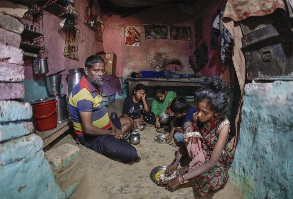 In this April 5, 2020, photo, Rajesh Dhaikar sits in his one room small house as his family eats dinner in Prayagraj, India. Dhaikar has a small balloon stall in a nearby market, selling plastic bursts of red and blue and yellow one at a time, and rarely earning more than $2.50 a day. His wife, Suneeta, makes about $20 a month cleaning homes. They have five children, ranging in age and a bank account with about $6.50 in it. India has launched one of the most draconian social experiments in human history, locking down its entire population, including hundreds of millions of people who struggle to survive on a few dollars a day. Image by AP Photo/Rajesh Kumar Singh. India, 2020.