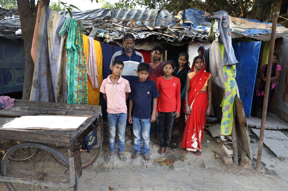 In this April 1, 2020, photo, Rajesh Dhaikar, rear left, stands with his family members, son Deepak and daughter Arti in the background and, from left in front row, sons Deepu, Raj, daughter Sapna and wife Suneeta, outside their one room house in Prayagraj, India. Dhaikar has a small balloon stall in a nearby market, selling plastic bursts of red and blue and yellow one at a time, and rarely earning more than $2.50 a day. His wife, Suneeta, makes about $20 a month cleaning homes. They have five children, ranging in age and a bank account with about $6.50 in it. India has launched one of the most draconian social experiments in human history, locking down its entire population, including hundreds of millions of people who struggle to survive on a few dollars a day. Image by AP Photo/Rajesh Kumar Singh. India, 2020.