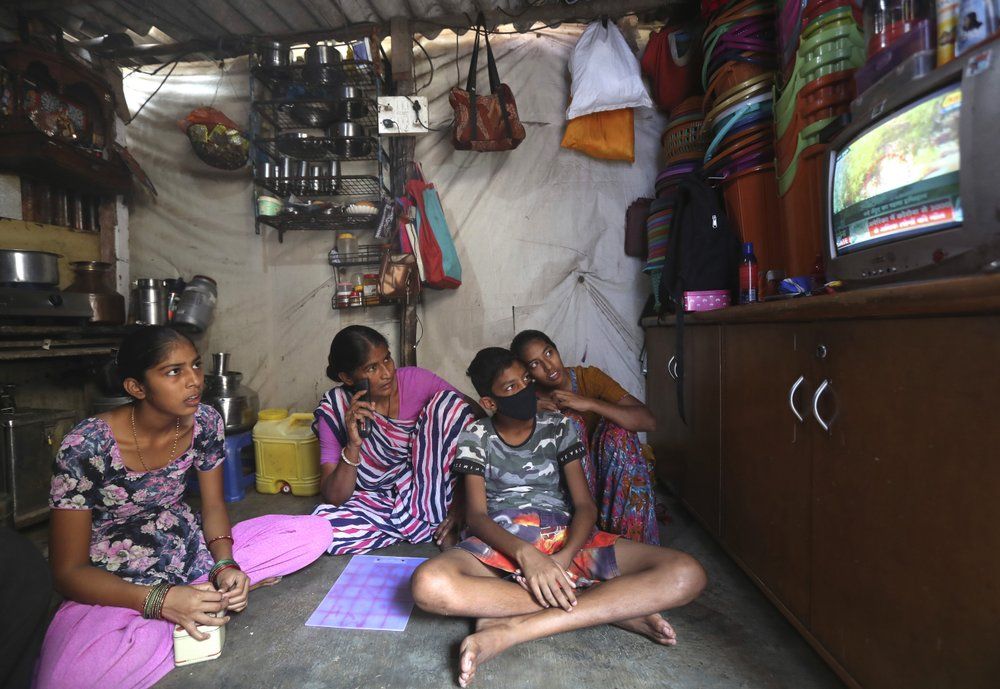 In this March 31, 2020, photo, Mina Ramesh Jakhawadiya, center, watches news on coronavirus along with her children in her one room house in a slum in Mumbai, India. Jakhawadiya makes a living selling cheap plastic goods with her husband on the streets of Mumbai. For her, the order means 21 days in a 6-by-9 foot room with five people, no work, a couple days of food and very less cash. As governments around the world debate ways to slow the spread of coronavirus, India has launched one of the most draconian social experiments in human history, locking down its entire population, including hundreds of millions of people who struggle to survive on a few dollars a day. Image by AP Photo/Rajesh Kumar Singh. India, 2020.