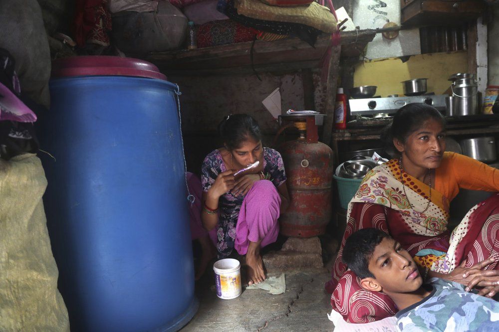 In this March 31, 2020, photo, Mina Ramesh Jakhawadiya, right with her son Ritik Ramesh in her lap watches news on coronavirus as her daughter Guddi Ramesh brushes her teeth in their one room house in a slum in Mumbai, India. Jakhawadiya, who makes a living selling cheap plastic goods with her husband on the streets of Mumbai. For her, the order means 21 days in a 6-by-9 foot room with five people, no work, a couple days of food and very less cash. As governments around the world debate ways to slow the spread of coronavirus, India has launched one of the most draconian social experiments in human history, locking down its entire population, including hundreds of millions of people who struggle to survive on a few dollars a day. Image by AP Photo/Rajesh Kumar Singh. India, 2020.