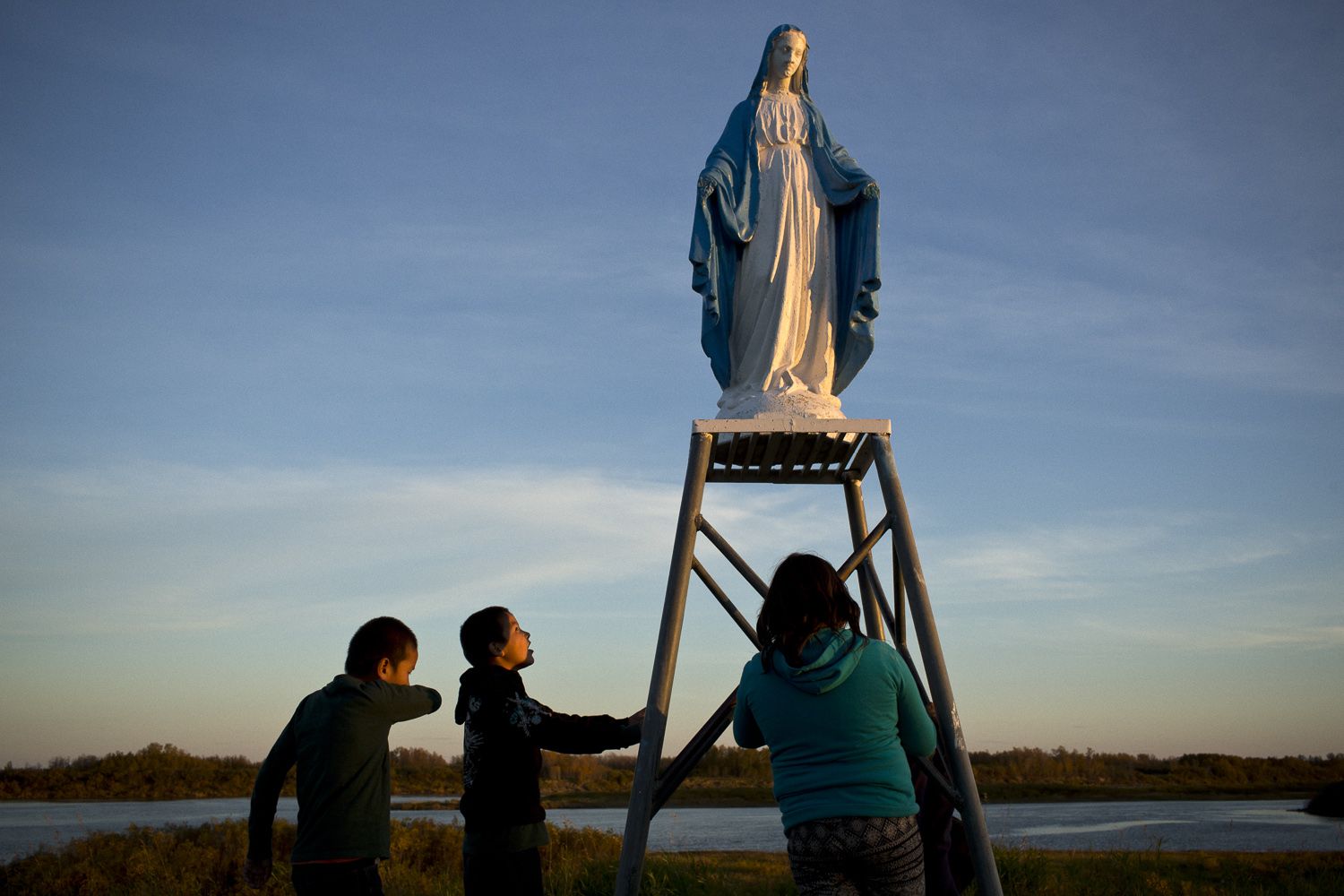 Young people play at a statue of the Virgin Mary near the St. François-Xavier Catholic Church on the shores of the Attawapiskat River in northern Ontario. The community has a complicated history with the Catholic Church, with many of its members subjected to horrible abuses while at the Catholic-run St. Anne’s Residential School. Image by David Maurice Smith/Oculi. Canada, 2016.