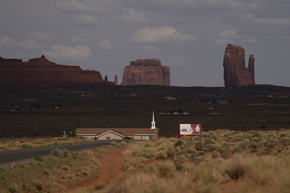 The mesas of Monument Valley are seen beyond the Church of Jesus Christ of Latter-day Saints in Oljato-Monument Valley, Utah on the Navajo Reservation April 30, 2020. The reservation has some of the highest rates of coronavirus in the country. If Navajos are susceptible to the virus' spread in part because they are so closely knit, that's also how many believe they will beat it. Image by AP Photo/Carolyn Kaster. United States, 2020.