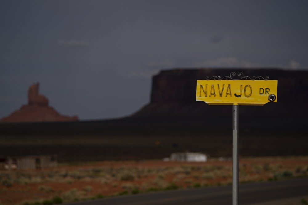 A sign for Navajo Drive is seen against a cloud-darkened Sentinel Mesa in Oljato-Monument Valley, Utah on the Navajo Reservation on April 30, 2020. The reservation has some of the highest rates of coronavirus in the country. If Navajos are susceptible to the virus' spread in part because they are so closely knit, that's also how many believe they will beat it. Image by AP Photo/Carolyn Kaster. United States, 2020.