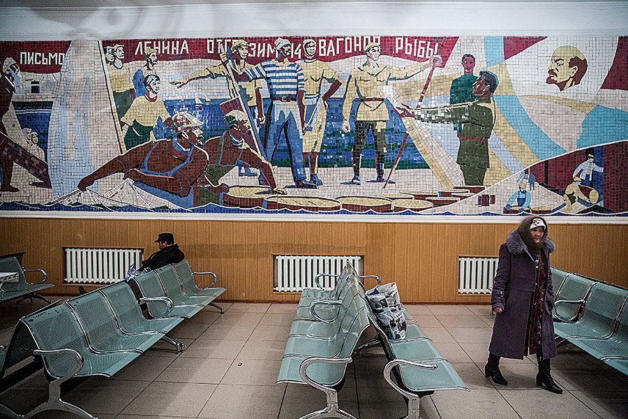 A Kazakh man and woman wait for a train under a Soviet mural in Aralsk. Kazakhstan declared independence from the Soviet Union in 1991. The mural commemorates the events during the 1921 famine when Vladimir Lenin wrote a letter to Aralsk asking for assistance from the fishermen. They sent 14 rail wagons of fish to the famine-ravaged regions. Image by Taylor Weidman. Kazakhstan, 2018.