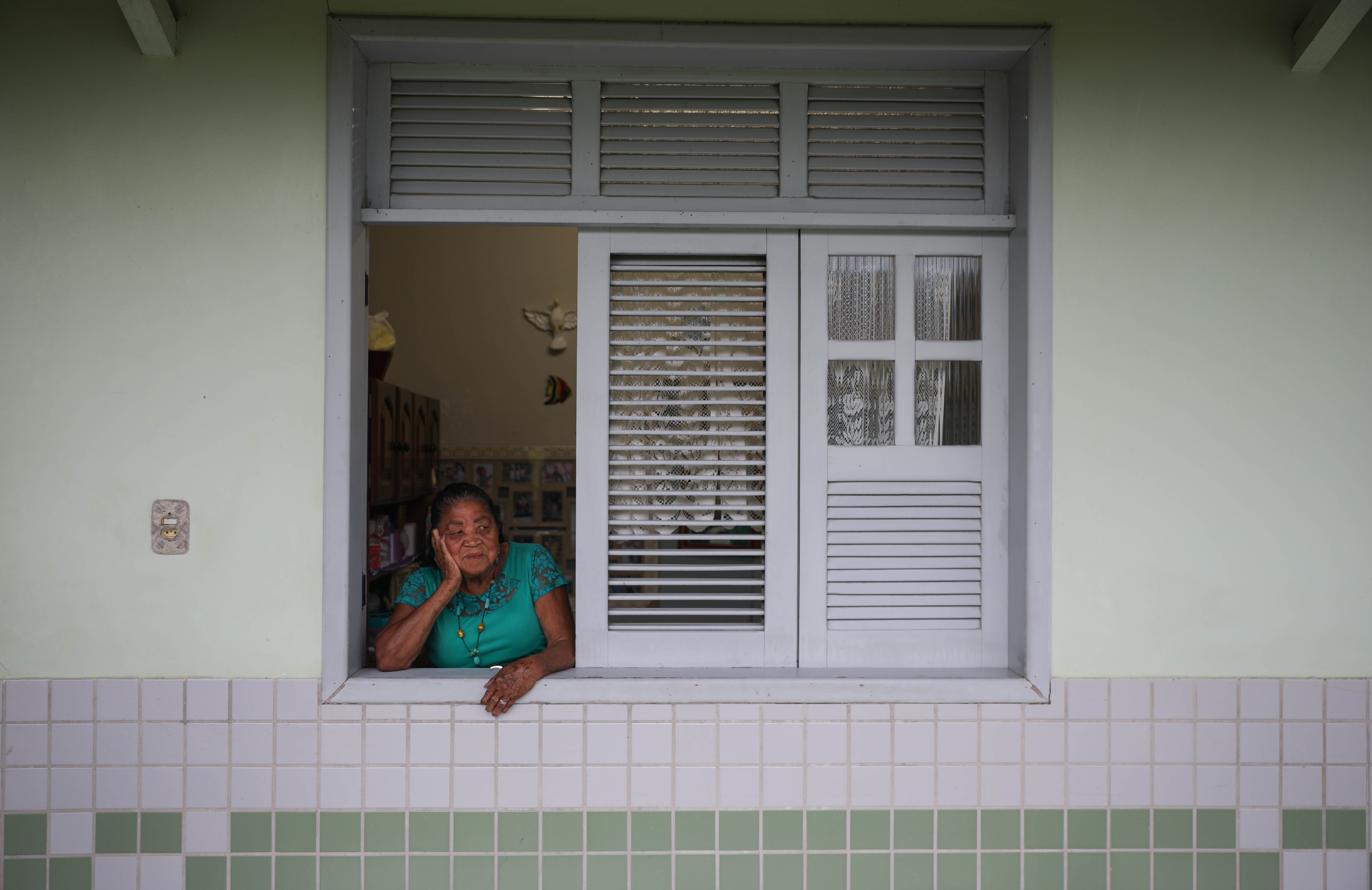 Maria da Silva Trindade peeks out of her living room window to chat with the other residents of Marituba’s former leprosy colony. Trindade was diagnosed with the disease at the age of 15, and she moved into the colony the same year. Nearly 80 years later she’s still there. Image by Anton L. Delgado. Brazil, 2020.