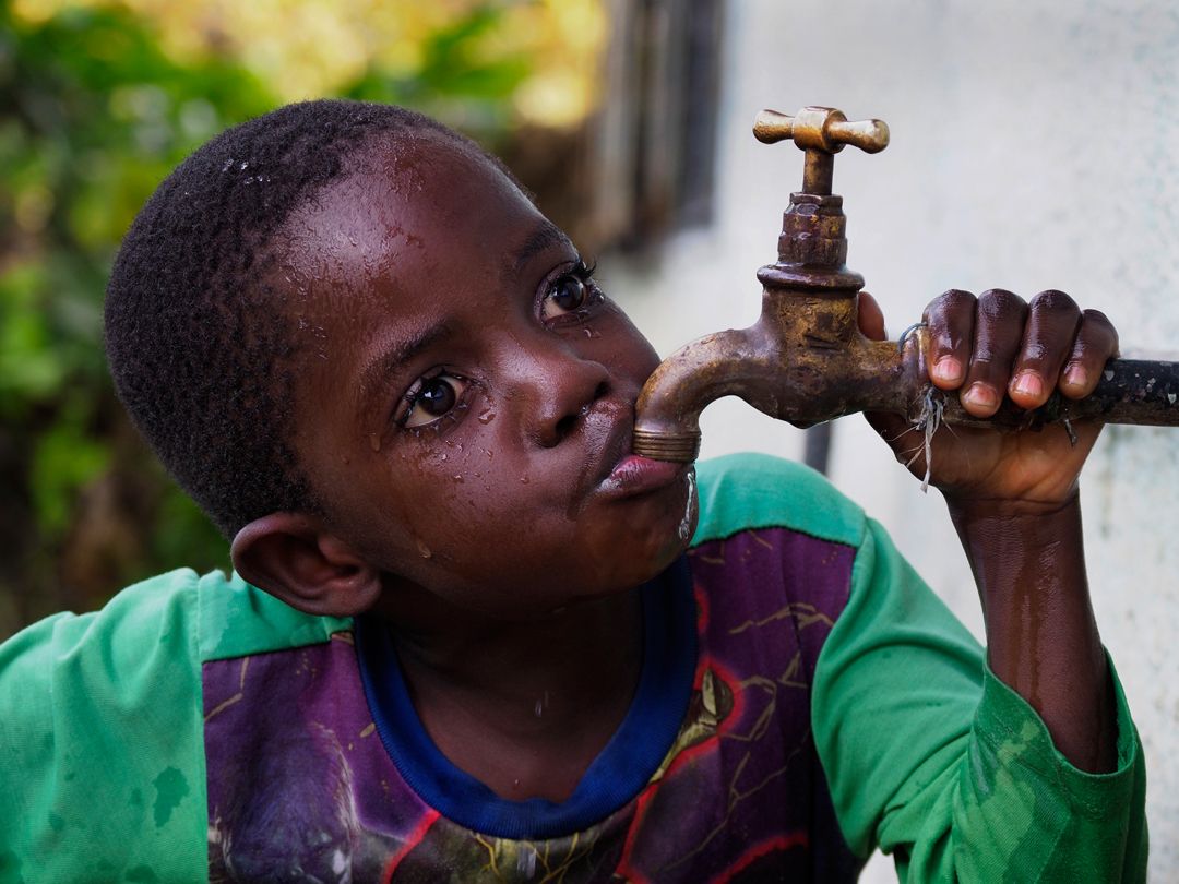 Lead poisoning victim: Gift Phiri, 6, drinks from a faucet in his backyard in Chowa, the heavily polluted neighborhood next to the closed lead mine. Phiri tested positive in 2015 for high blood lead levels. Image by Larry C. Price. Zambia, 2017.