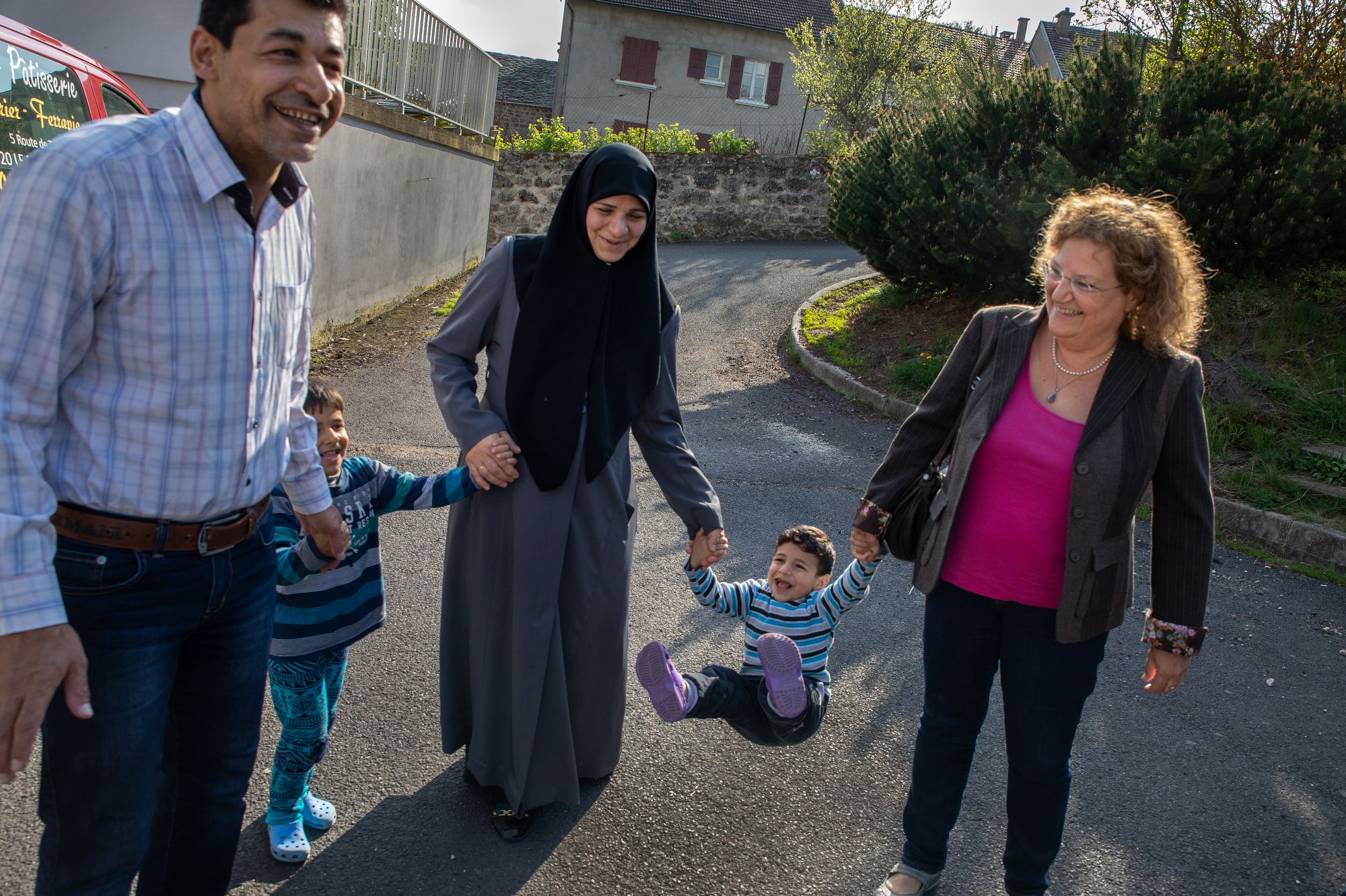 Family members of Marianne Bouvier-Mermet (far right), were honored for hiding Jewish refugees during World War II. She carries on that tradition by working with refugees including this Syrian family of Ahmed, his pregnant wife, Ibtesam, and their two children, Mohamed-Nour (5) and Abdurrhaman (3).   Bouvier-Mermet has helped them since their arrival last winter, including letting them live in an apartment she owns. After the horror the Syrian family faced in their hometown of Alleppo and spending several years in a Turkish refugee camp, they are happy with their life in Mazet-Saint-Voy, “Everybody here says bonjour to you,” explains Ahmed. For Marianne helping them and other refugee families is part of the history of the Plateau. “For us it is necessary that this past serve the present, and that the reception of the humble inhabitants of the Plateau is an example for now.” She said. Image by Lucian Perkins. France, 2018.