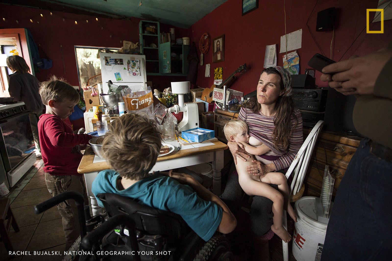 Allison, a mother of eight, breastfeeds her youngest daughter at the kitchen table in their two-bedroom cabin on a commune in the redwood forest. Past a gate marked ‘Private Property — Keep Out,’ the Murphy family live with no plumbing, indoor bathroom, heat, or central air. Image by Rachel Bujalski. United States.