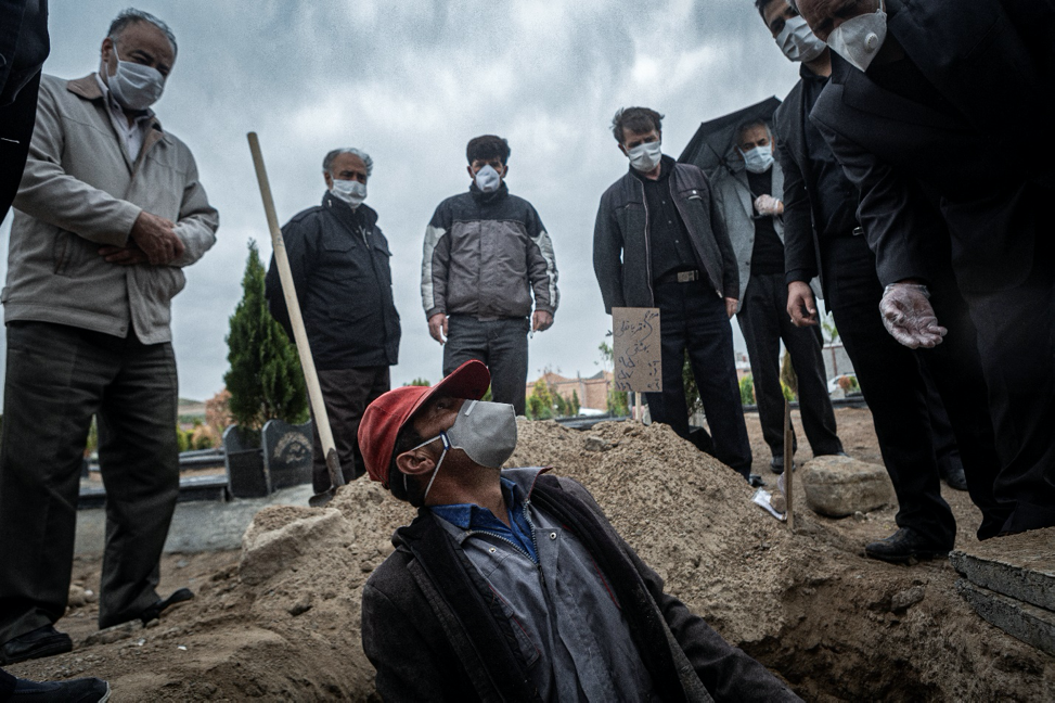 Tabriz, Iran, May 6, 2020. The worker has put the corpse inside the grave while having a mask on his face and he asks the dead man’s relatives to take some distance, thus he can provide the deceased an Islamic burial ceremony. Iran was amongst five of the countries with the highest spread of COVID-19. Iran’s government began an official lockdown on March 15, 2020, the quarantines in place are for those who do not exhibit symptoms but have been exposed to the illness. For the large number of Iranians who live on a day-by-day income, staying home means having no money to pay for essential things such as food. Therefore, they must take their life in their own hands and work in order to survive even at the risk of getting infected, spreading the virus in society at large, and even getting fined for breaking the quarantine. For them, choosing either path has extreme consequences.  Image by Jalal Shamsazaran / NVP Images. Iran, 2020.