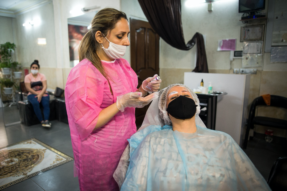 Tehran, Iran – April, 2020. Manijeh Kamandloo, 43, from Tehran, working as a beauty specialist under heavy protection during the coronavirus pandemic. She has been a hairdresser for almost 20 years and only recently ran a beauty salon of her own in the east of Tehran. She is a mother of two and the sole provider for her family. She had an income of about eight million Iranian tomans ($470) before the outbreak of the coronavirus disease, but since March she has been made to close her salon for public safety. Nowadays she is back to work again, however, the number of her customers has significantly decreased causing her income to be near a shocking amount of 2 million Iranian tomans. “I’m spending most of my earnings on disinfectants, masks, and clothes, thereby almost little money will be left at the end of the month,” she said.  Experiencing a financially tough time, she is thinking of closing her salon. Image by Yasaman Dehmiyani / NVP Images. Iran, 2020.