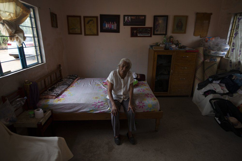Catalina Vazquez poses for a portrait as she sits on her bed in San Jeronimo Xayacatlan, a town in Mexico from where nearly a third have emigrated to New York, Thursday, June 25, 2020. Vazquez's daughter Magnolia Ortega works cleaning homes in New York, and her son Jorge Vazquez returned from New York in the early 2000s after emigrating twice. Here, she and Jorge care for Magnolia's three daughters, with the help of money she sends home. In total, three of Catalina's children are in the U.S. and two are in Mexico. Image by Fernando Llano/AP Photo. Mexico, 2020.
