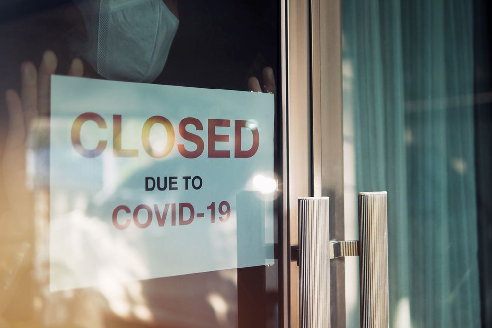 Business office or store shop is closed, bankrupt business due to the effect of novel Coronavirus (COVID-19) pandemic. Unidentified person wearing mask hanging closed sign in background on front door. Image by Chansom Pantip. Thailand, 2020.