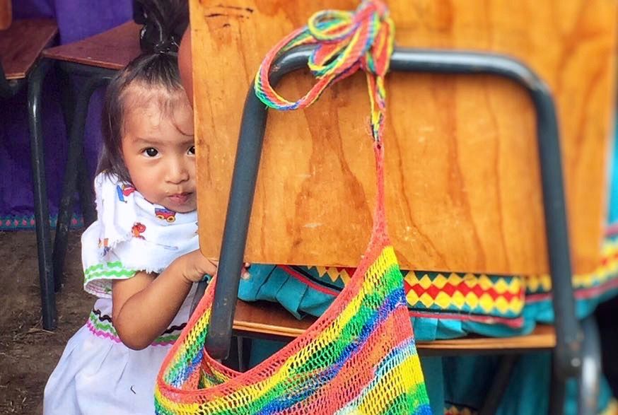 A young Ngäbe-Buglé girl sneaks into a "chácara," a traditional woven bag, latched to her mother’s chair during a community cultural event. Image by Samira Tella. Costa Rica, 2018. 