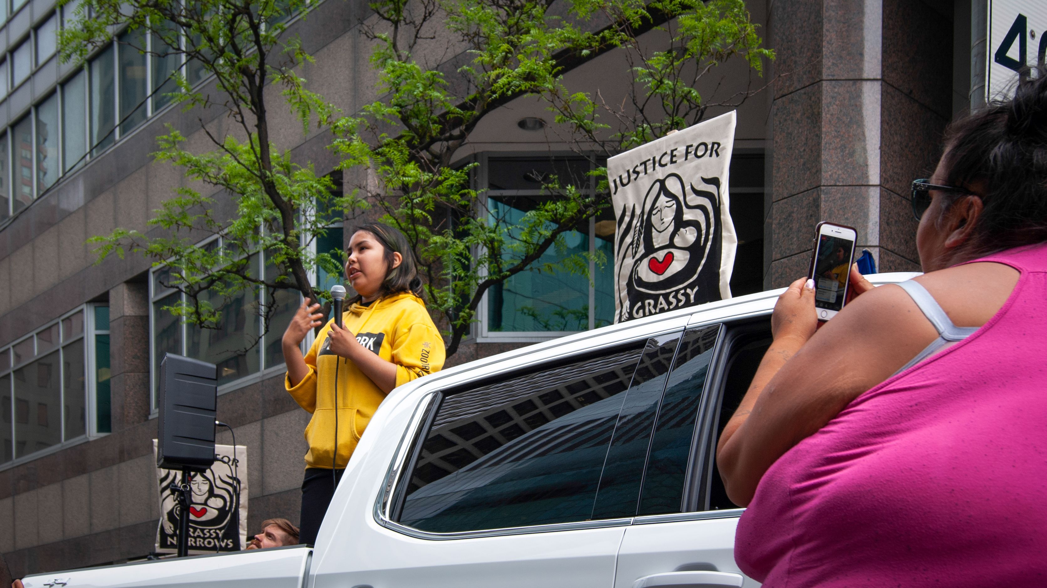 Jenae Turtle (left) stands before a crowd and speaks about her community while Chrissy Isaacs (right) stands below her, filming Jenae's speech. Canada, 2019. Image by Shelby Gilson.