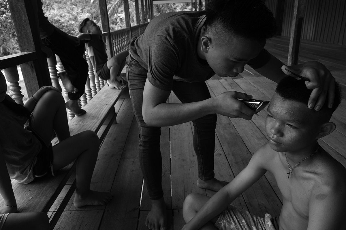 One Penan boy cuts another's hair on the veranda of their longhouse, Long Jaik. Long Jaik is in the midst of a legal land dispute iwth a company that sells timber and palm oil. Image by Stuart Franklin/Magnum Photos. Malaysia, 2018.