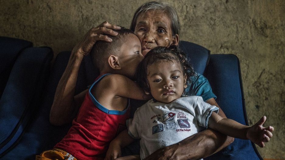 Remy Fernandez, 84 years old, holds two of the seven grandchildren she is raising. Her youngest son, Constantino de Juan, a methamphetamine user, was killed by masked men in December, 2016. Upon seeing his attackers, Juan instructed five-year-old CJ, shown here wearing a red tank top, to take care of his siblings. The children's mother is in prison due to a drug arrest. Baby RJ, in the "daddy's little helper" T-shirt, was born in prison. Image by James Whitlow Delano. Philippines, 2017. 