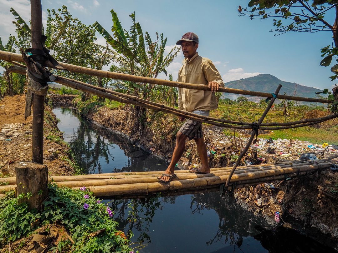 A farmer walks over a small bridge to cross an irrigation canal that feeds black waste water into rice fields near Rancaekek. Image by Larry C. Price. Indonesia, 2016.