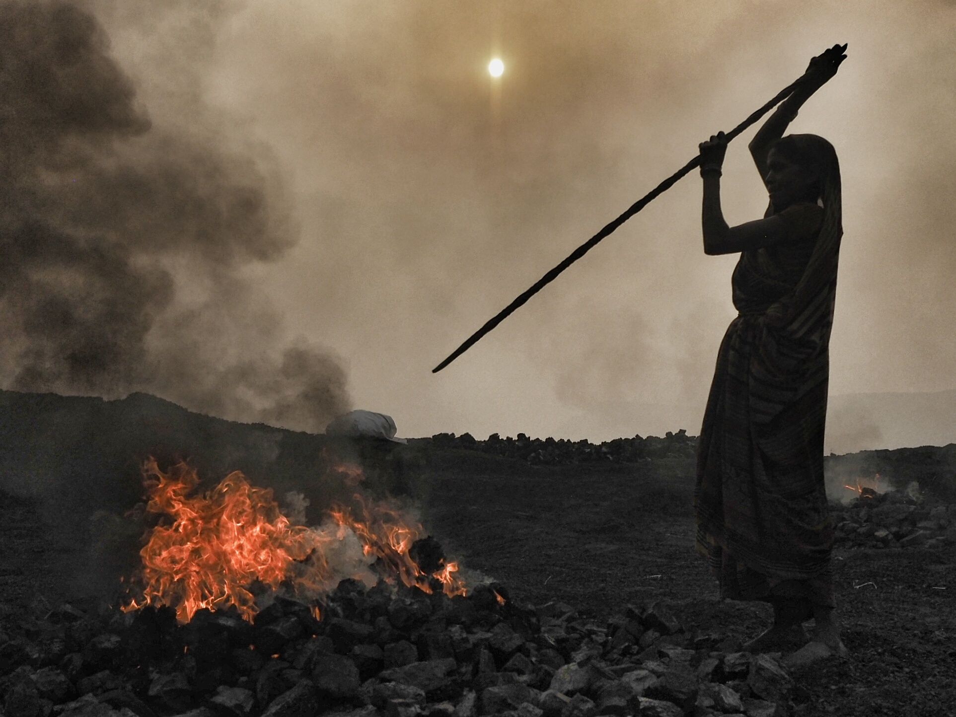 At sunrise, a woman jabs a steel into a burning pile of coal.