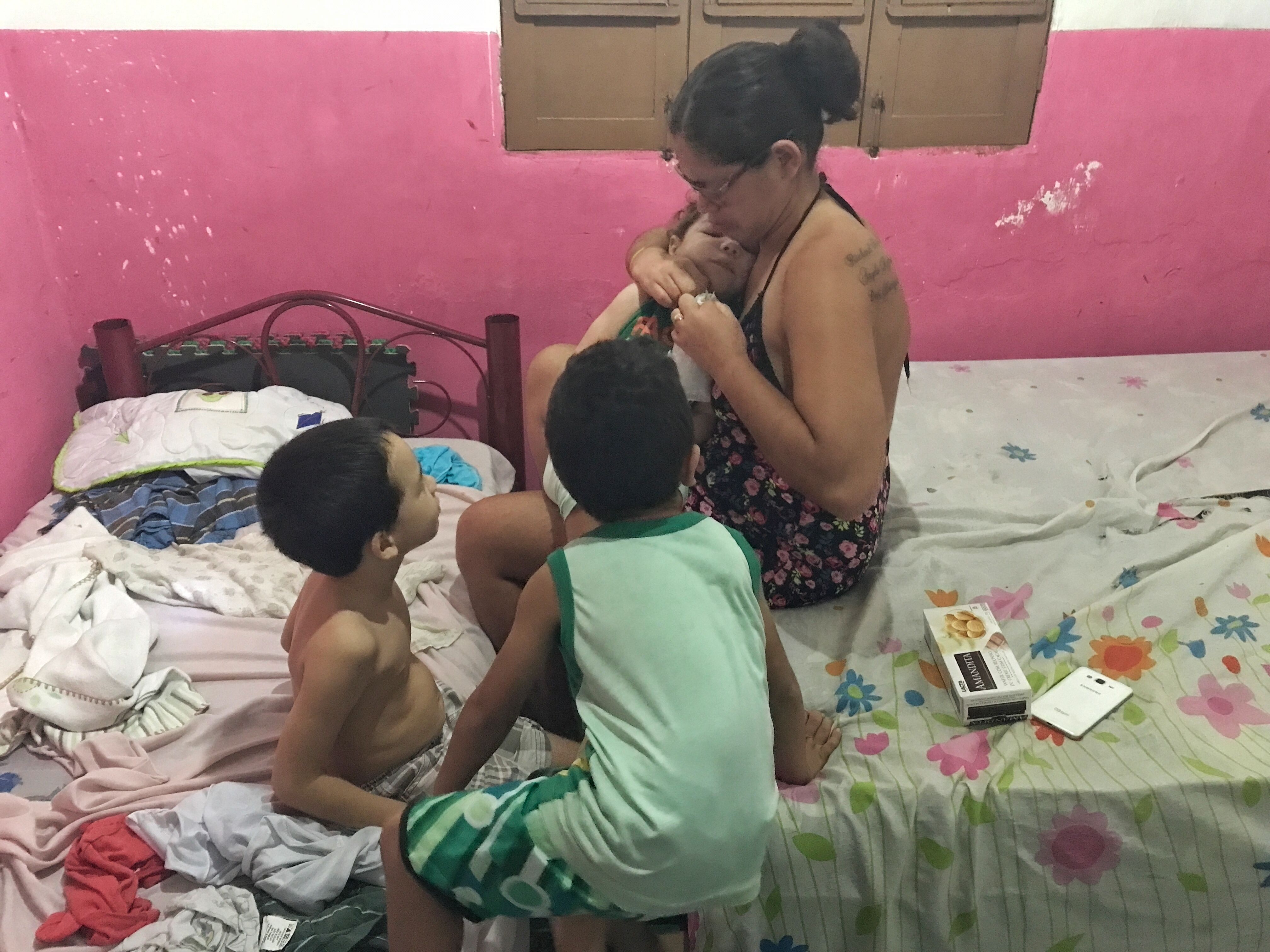 Mylene Helena dos Santos Ferreira, 23, opens a package of chocolate biscuits for her eager sons while balancing Davi in her lap. Davi Herrique Ferreira da Silva was born in August 2015 with congenital Zika syndrome. Shirtless on the twin bed is Mylene’s oldest son, five-year-old Richard Miguel; next to him in the green outfit is Angelo Rafael, age four. When Davi was only a few months old, Mylene left her abusive husband of eight years. He now refuses to acknowledge Davi, but when Davi falls sick, Mylene must send her older sons to stay with him. In July, Mylene was at the hospital with Davi for nearly two weeks while he was treated for a severe respiratory infection. Miguel and Rafael did not see her during this time. “Whenever I talked to them on the phone, they asked when I was coming home,” Mylene said. These pictures were taken the week she and Davi returned from the hospital. Image by Poonam Daryani. Brazil, 2017.