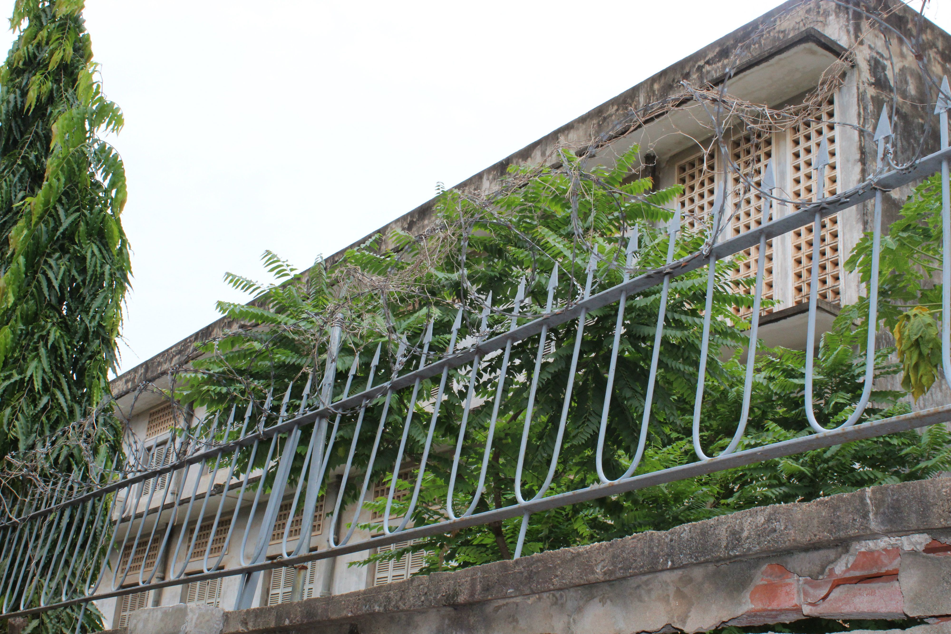 Photograph of S-21 former concentration camp, taken from the perimeter wall.  Barbed wires, spiked metal railings, and overgrown ferns partly shield the view of the building. Image by M.G. Zimeta. Cambodia, 2018.