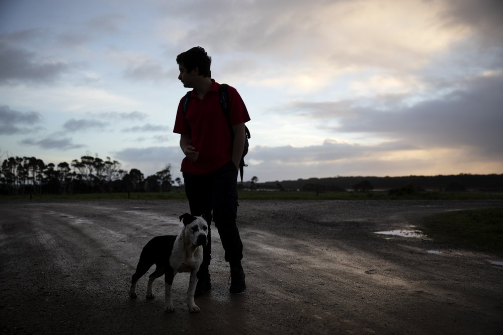 CORRECTS LAST NAME - Joe Watkins, 15, waits with his dog, Ellie, for the school bus near his home in Black River, Tasmania, Australia, Wednesday, July 24, 2019. Watkins’ mother was a dairy farmer when her drug addiction all began, a cheerful woman who loved taking Joe fishing and watching her daughter, Sarah, ride horses. And then one day, ten years ago, she jumped off a truck and felt her knees give way. Image by David Goldman. Australia, 2019.