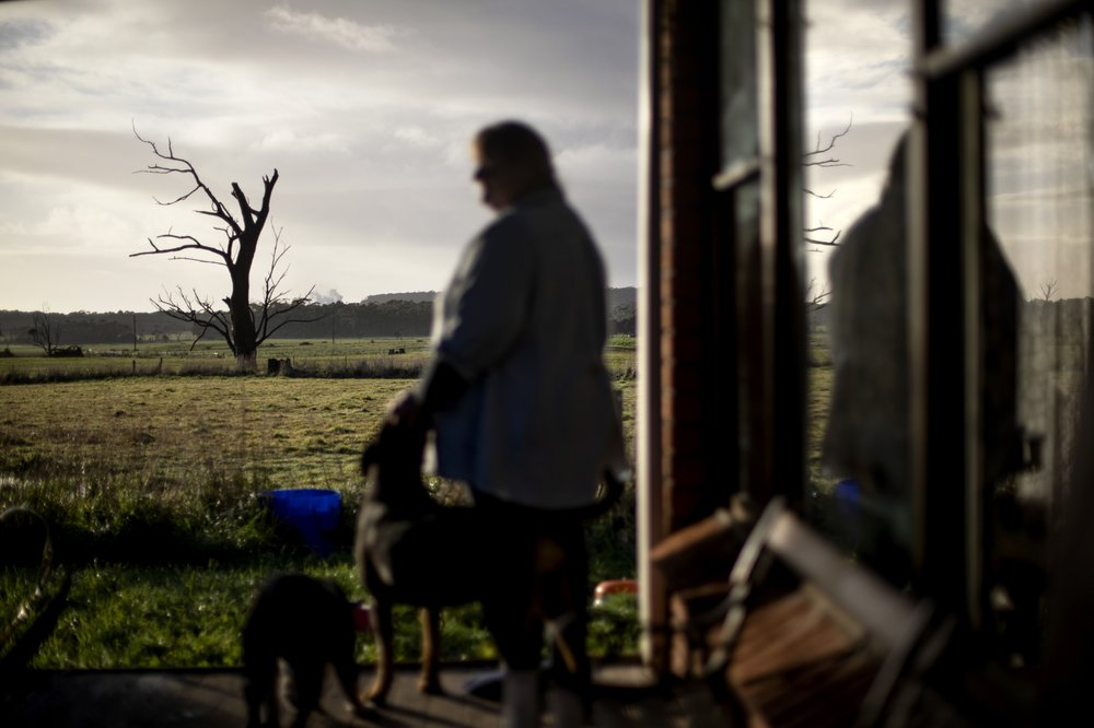 With her favorite tree in the background, Carmall Casey stands for a photo on her front porch in Black River, Tasmania, Australia, Wednesday, July 24, 2019. The tree is dead and gnarled, but still standing. Like her, she says, it's old and broken, but still good for something. "But dead trees, even though they're dead, they've still got beauty of some kind haven't they?" Image by David Goldman. Australia, 2019.