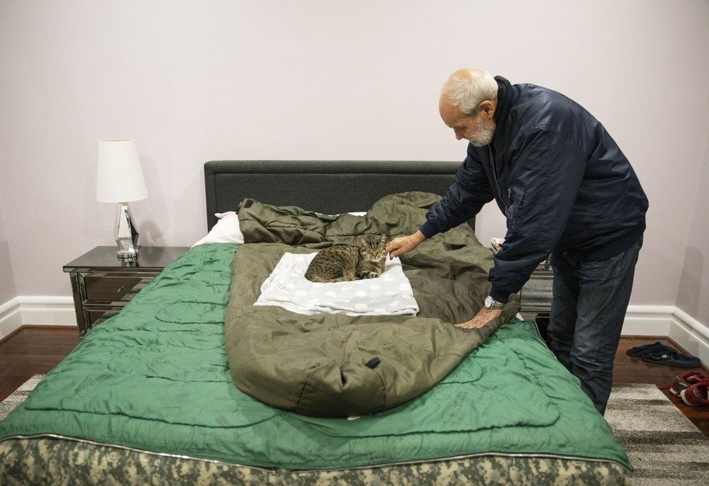 David Tonkin pets Ned, his son Matthew's cat, that now sleeps on David's bed on the sleeping bag Matthew used in Afghanistan, in Perth, Australia, Sunday, July 21, 2019. After Matthew died, and for years to come, David would suddenly awaken at 10 p.m. _ the same time that Matthew used to call from Afghanistan. Now, instead of Matthew's voice, there is only silence. Just like the silence from the officials he believes did little to prevent the death of his son. Image by David Goldman. Australia, 2019.