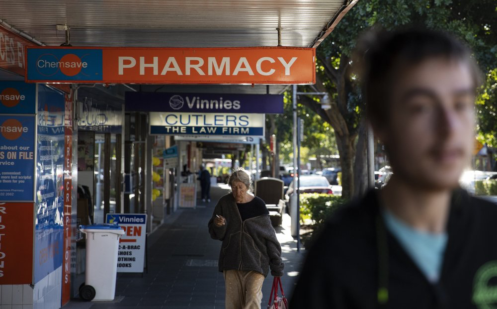 Sam Ware, 22, right, walks by the pharmacy where he used to get some of his opioid prescriptions filled in The Entrance, Central Coast, Australia, Thursday, July 25, 2019. The drugs were cheap. Most medications in Australia are government subsidized. For people like Sam who have concession cards _ those who are older, or on low incomes, or have a disability _ the out-of-pocket cost is just 6.50 Australian dollars (US$4.50) per prescription. Image courtesy of David Goldman. Australia, 2019.