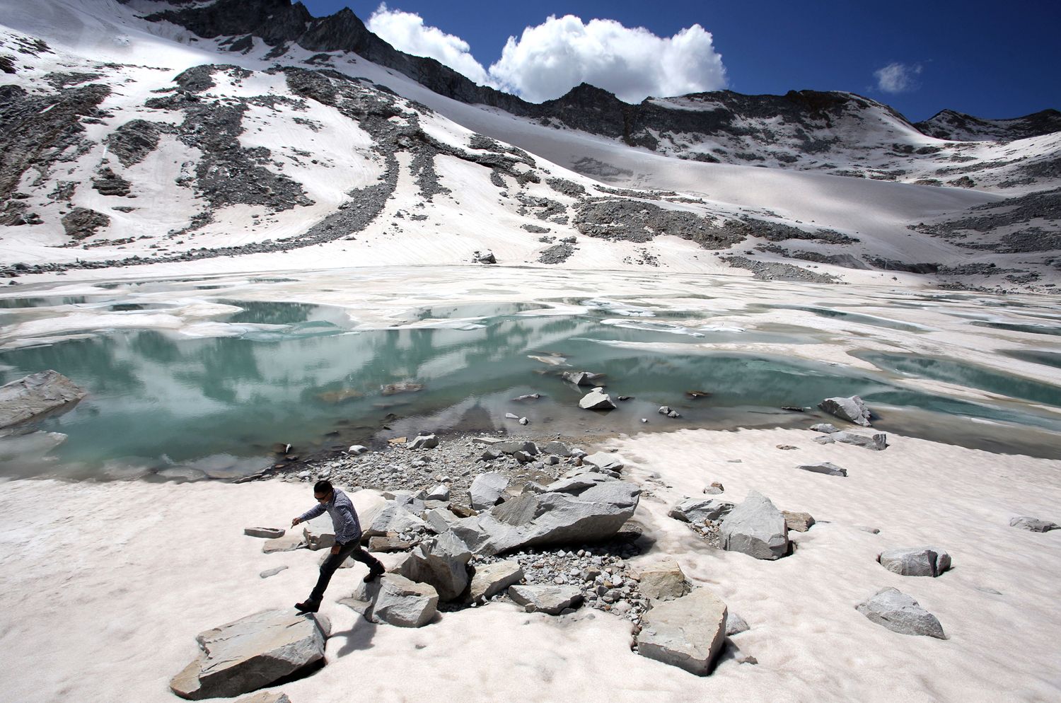 A man walks over rocks near to a glacial lake that has formed at the base of the Dagu Glacier on the southeast edge of the Tibetan Plateau. The glacier has been reducing in size in recent years as a result of rising temperatures in the region. Image by Sean Gallagher. China, 2012.