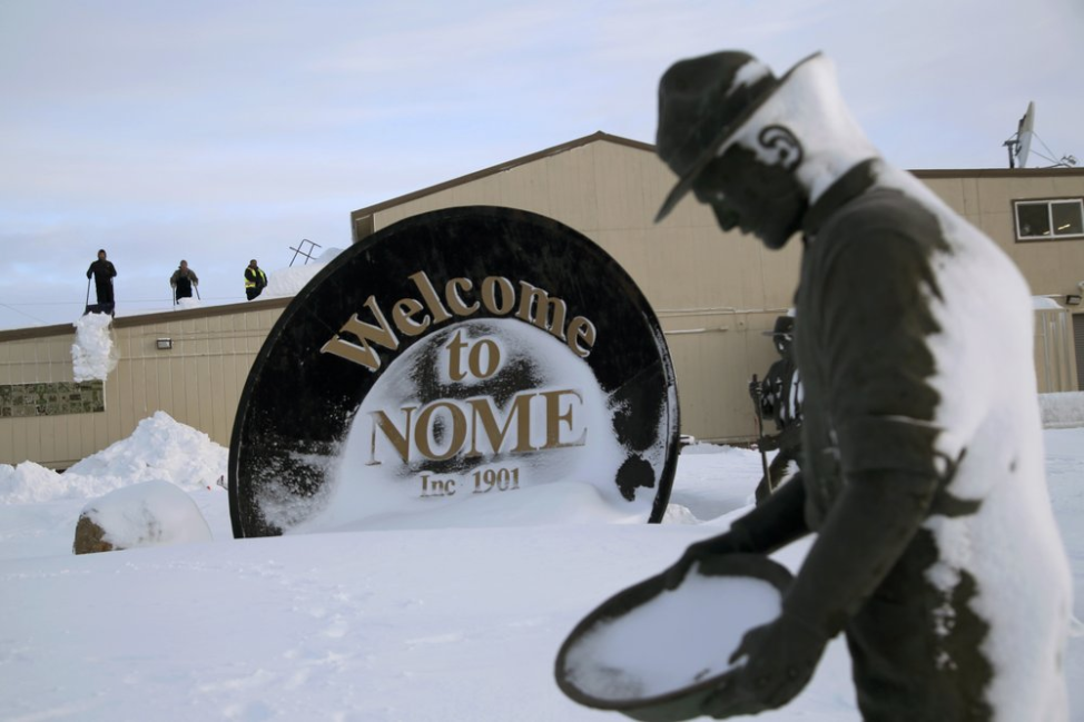 In this Feb. 14, 2019, photo, three men shovel snow from the roof of a grocery store as a statue of one of the "Three Lucky Swedes," credited with discovering gold in the late 19th century, stands in the foreground, in Nome, Alaska. The city later added statues of two native boys who led him to find the gold. Image by Wong Maye-E. United States, 2019.