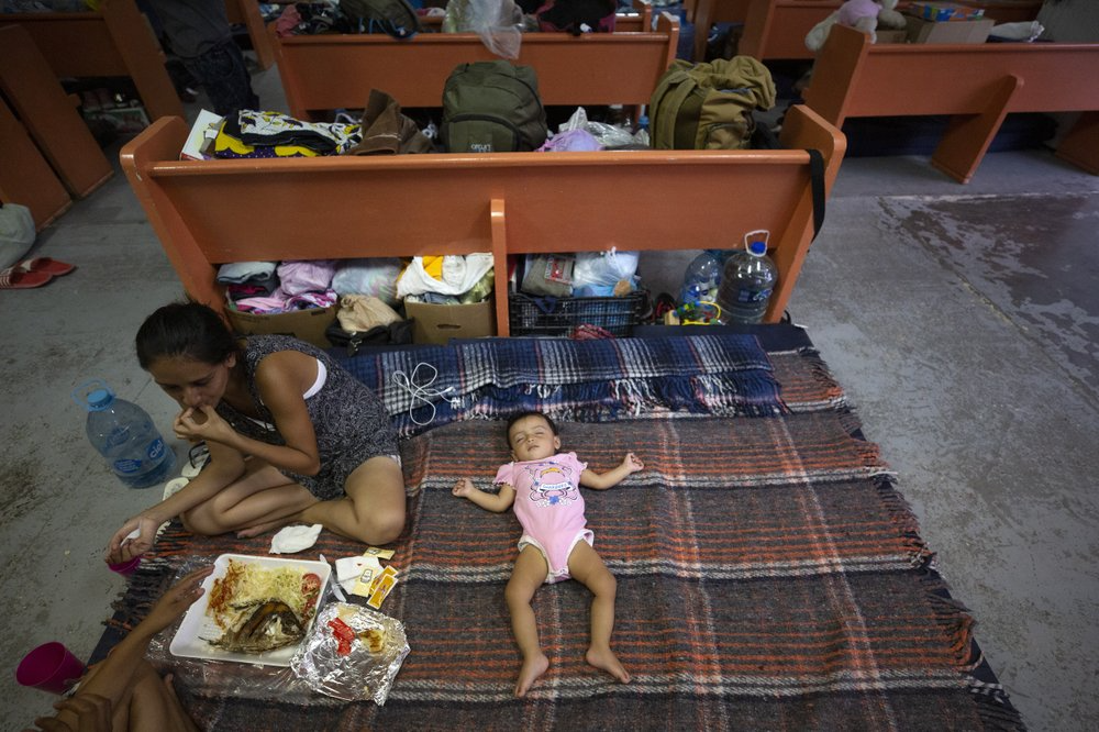 In this July 28, 2019, photo, Evelyn, of El Salvador, eats a shared lunch as her daughter sleeps in an area set up for families among the pews at El Buen Pastor shelter for migrants in Cuidad Juarez, Mexico. Image by Gregory Bull. Mexico, 2019.
