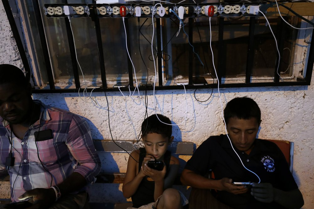 In this July 30, 2019, photo, migrants from Africa and Latin America check their phones among cables of charging phones plugged into sockets at El Buen Pastor shelter for migrants in Cuidad Juarez, Mexico. Image by Gregory Bull. Mexico, 2019.