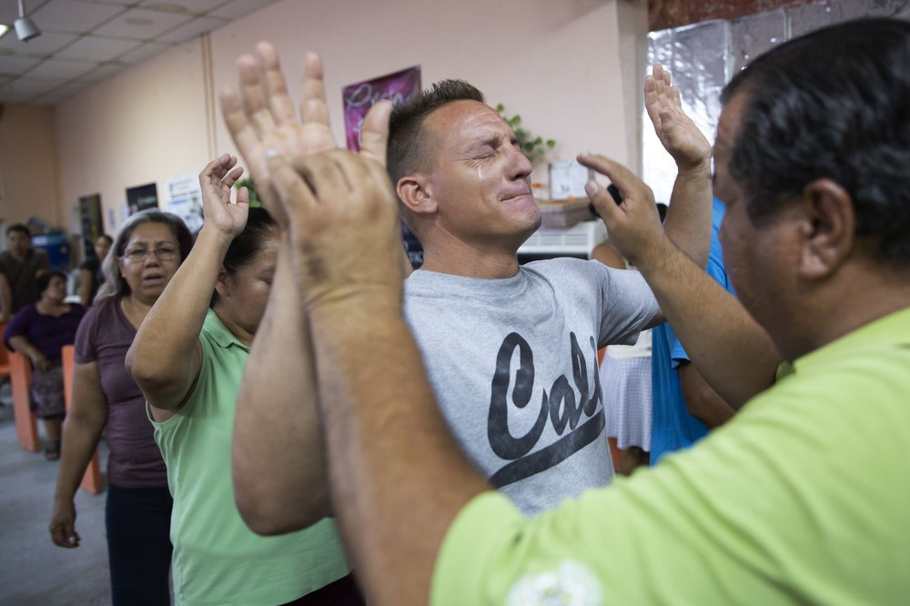 In this July 25, 2019, photo, pastor Juan Fierro, right, touches Miguel, of Cuba, during services at El Buen Pastor shelter for migrants in Cuidad Juarez, Mexico. Image by Gregory Bull. Mexico, 2019.