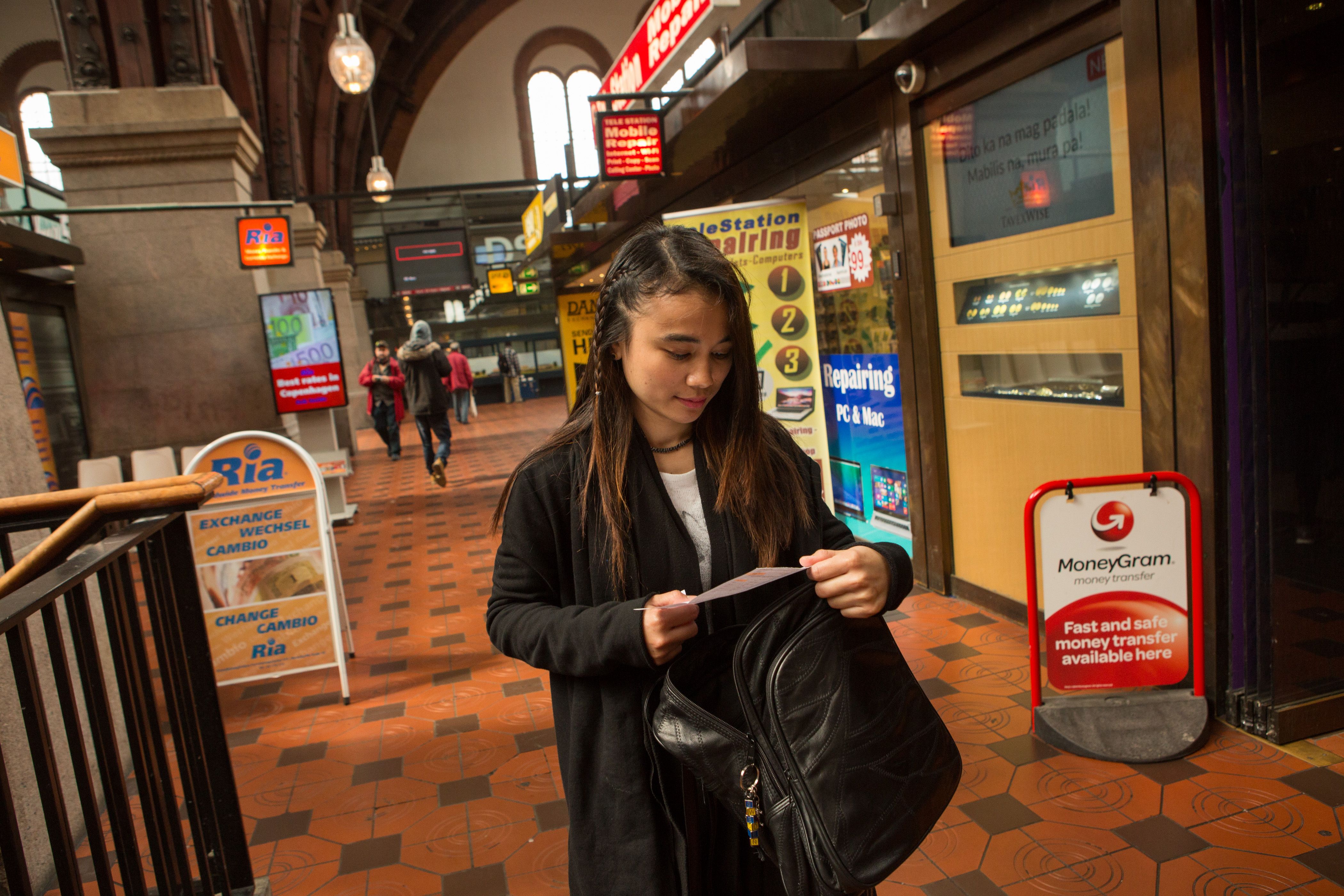 Au pair Lucila Espinosa Milay checks her receipt as she walks through a row of money transfer businesses after wiring part of her paycheck to siblings back in the Philippines, at Central Station in Copenhagen, Denmark, May 21, 2016. Milay pays the school fees for several of her eight siblings back home, sending money every 15 days. Remittances are a huge economic driver in the Philippines. Image by Allison Shelley. Philippines, 2016.