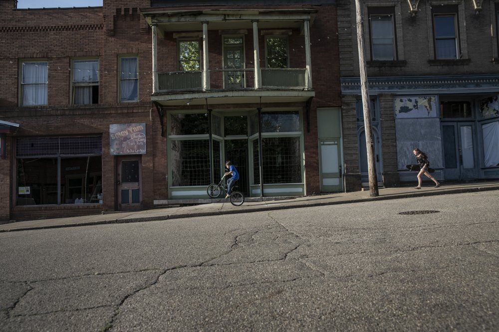 A boy rides his bicycle while a girl runs with her skateboard past abandoned storefronts along the streets of Shawnee, Ohio, on Friday, July 24, 2020. Shawnee was a coal town that once boasted an opera house, a vaudeville theater, dozens of stores and plenty of taverns. Today, Main Street is little but one abandoned building after another. Image by Wong Maye-E/AP Photo. United States, 2020.