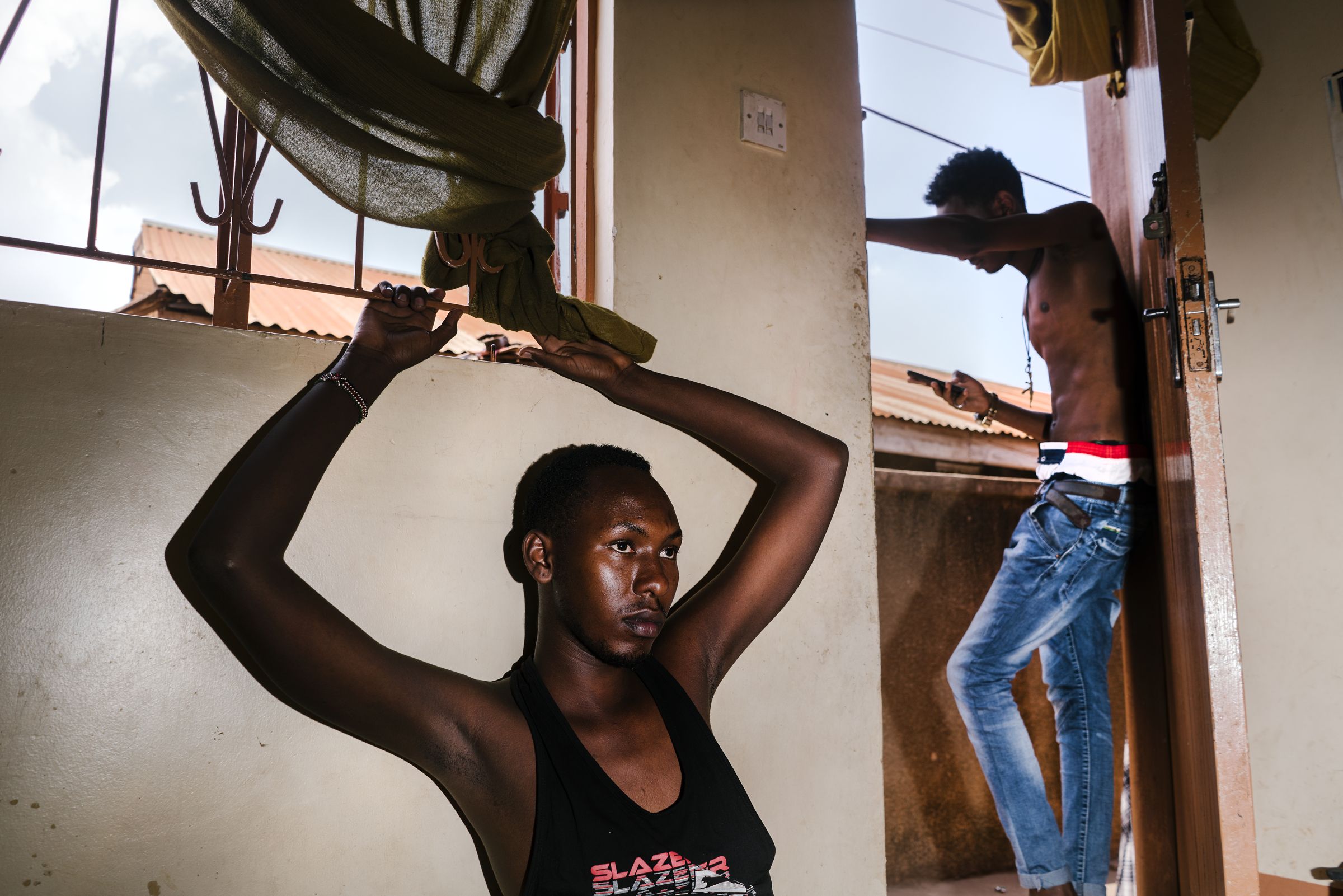 Javan sits in the family's living room, while her brother Shema stands in the doorway. Image by Jake Naughton. Uganda, 2017.