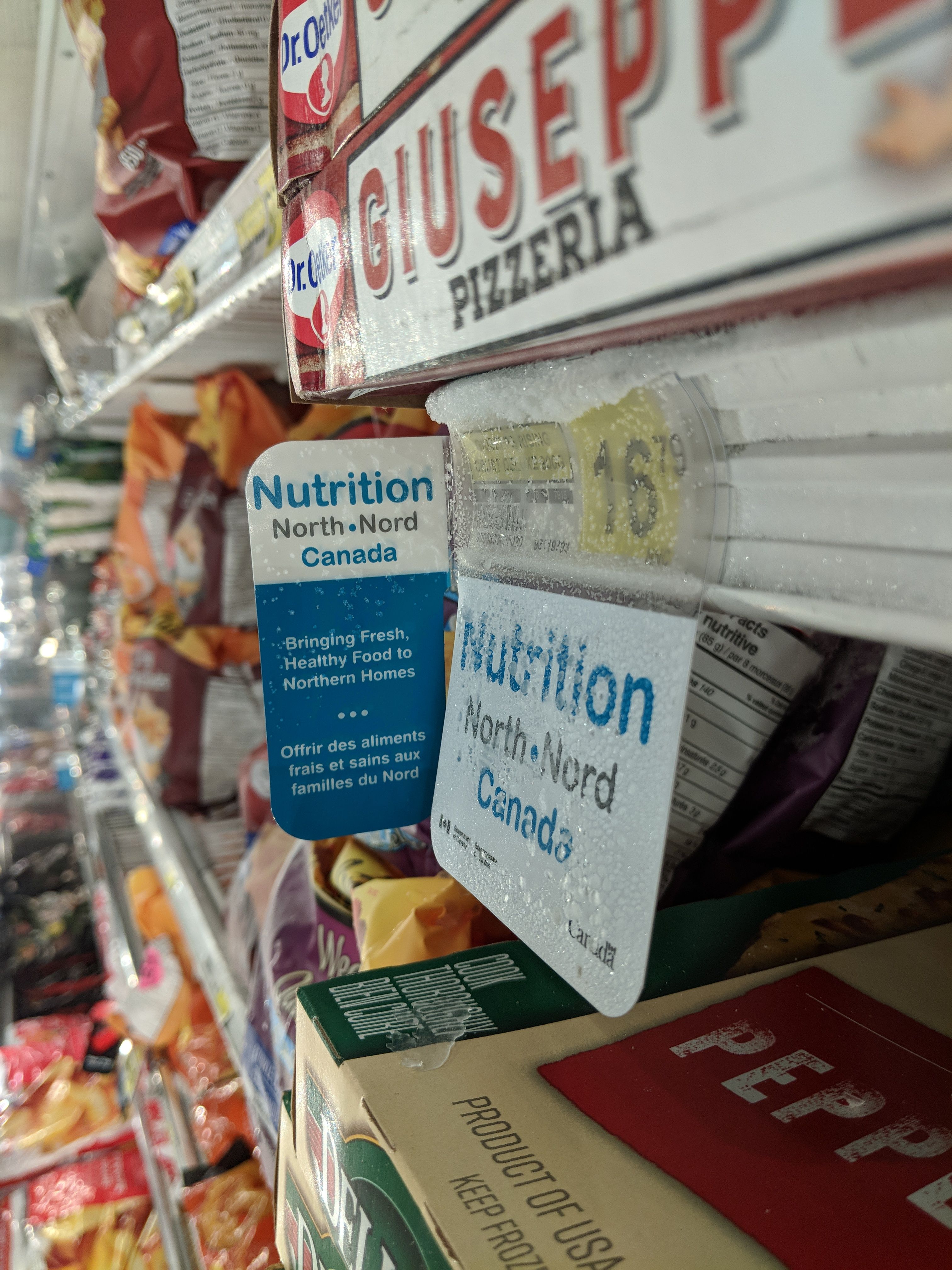 At a store in Pangnirtung, Nunavut, a sign for the Nutrition North program hangs above a frozen pizza (cost: $16.79). Inuit groups have been pressing Ottawa to reform the controversial subsidies for southern food flown into the North. Image by Julie De Meulemeester. Canada, 2018. 