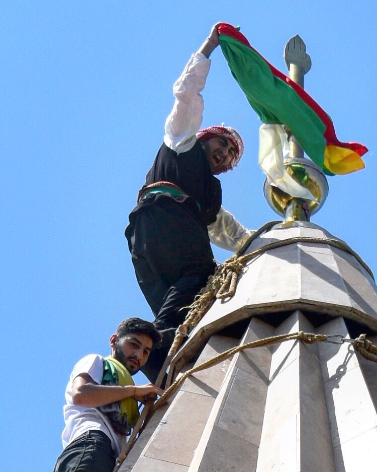After climbing the temple cupola, a young Yezidi man hoists a flag and waves it to cheers from the courtyard below. Image by Kaitlyn Johnson. Georgia, 2019.