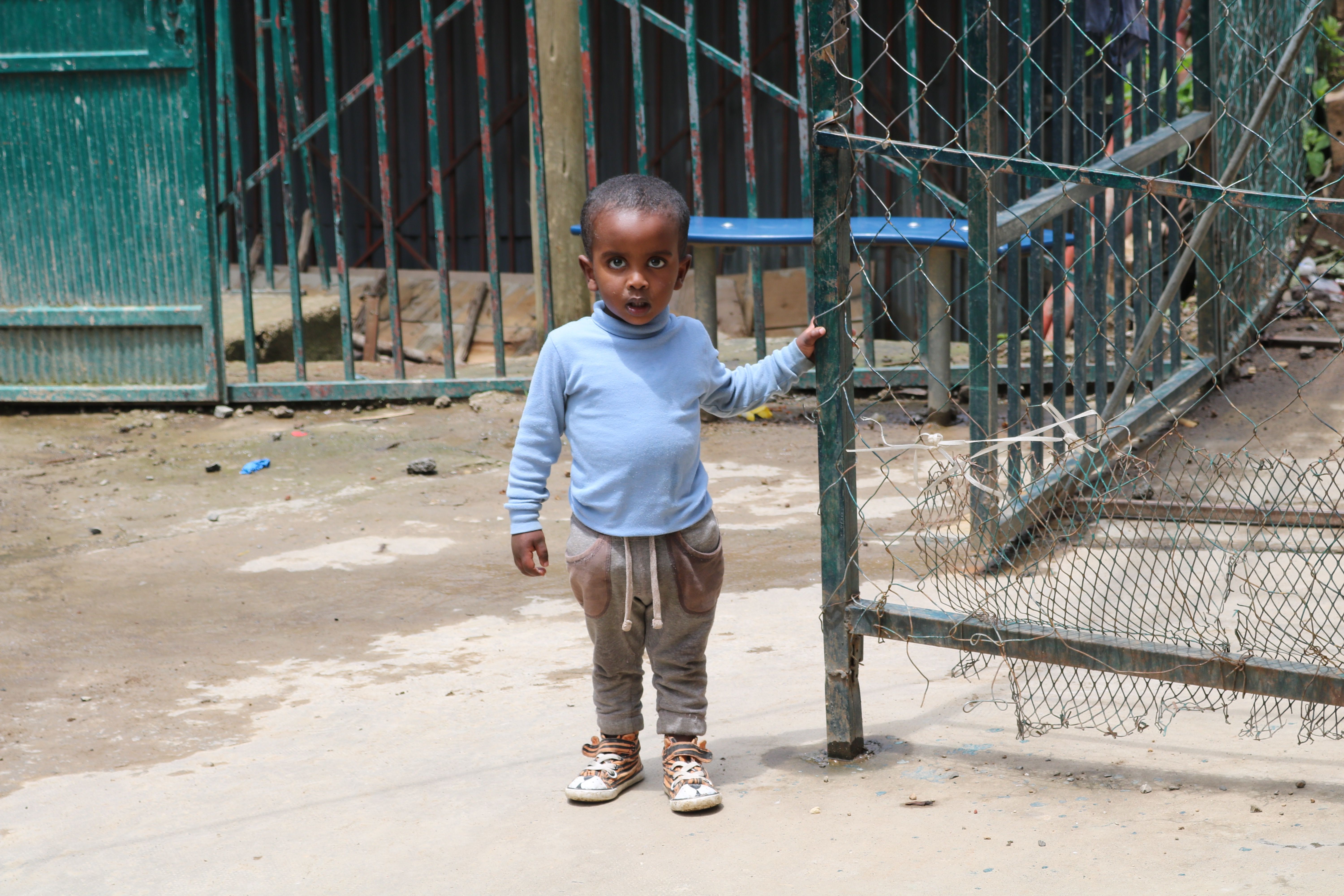 A young boy stands by the gate waiting to go inside to have lunch. He will spend his life in the orphanage unless he is locally adopted. Image by Abigail Bekele. Ethiopia, 2018.